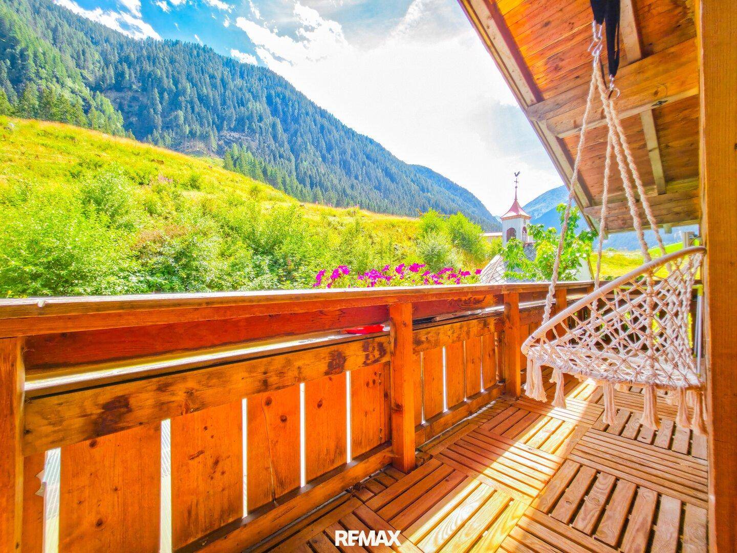 Sonniger Holzbalkon mit Hängesessel und weitem Blick auf die umliegende Berglandschaft.