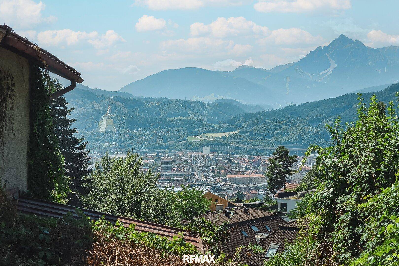 Weitläufiger Ausblick auf die Stadtlandschaft und die beeindruckende Bergkulisse unter blauem Himmel.