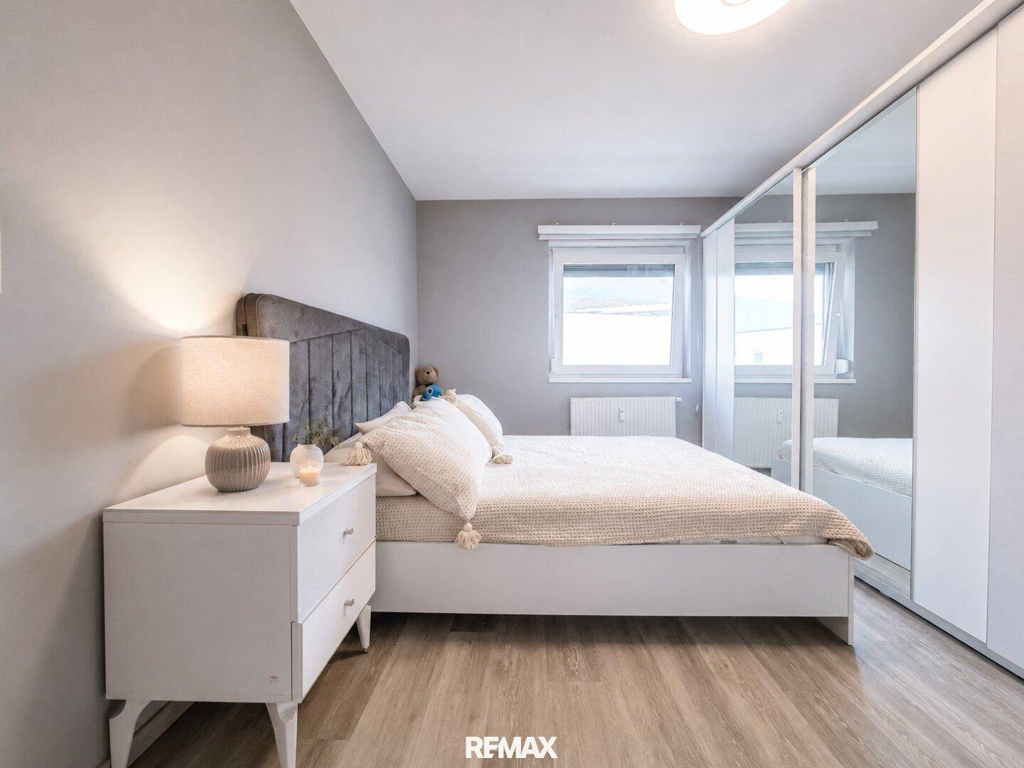 Gemütliches Schlafzimmer mit grauem Polsterbett, weißem Nachttisch und großem Spiegelschrank.