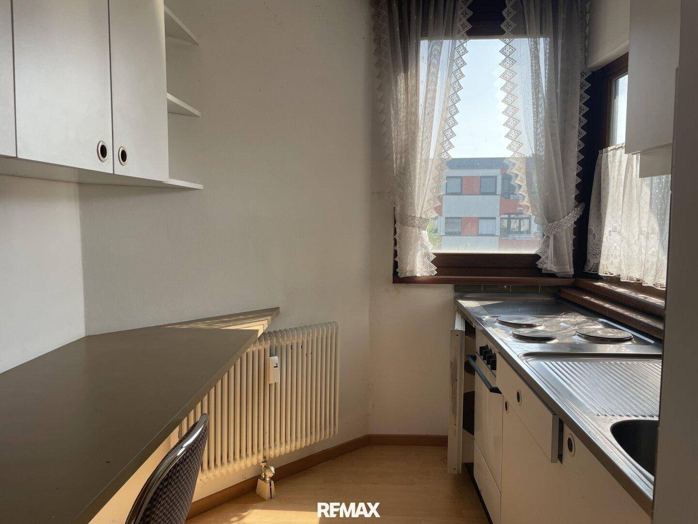 Kleine Küche mit weißen Oberschränken, Herd, Spüle und einem Fenster mit Vorhängen.