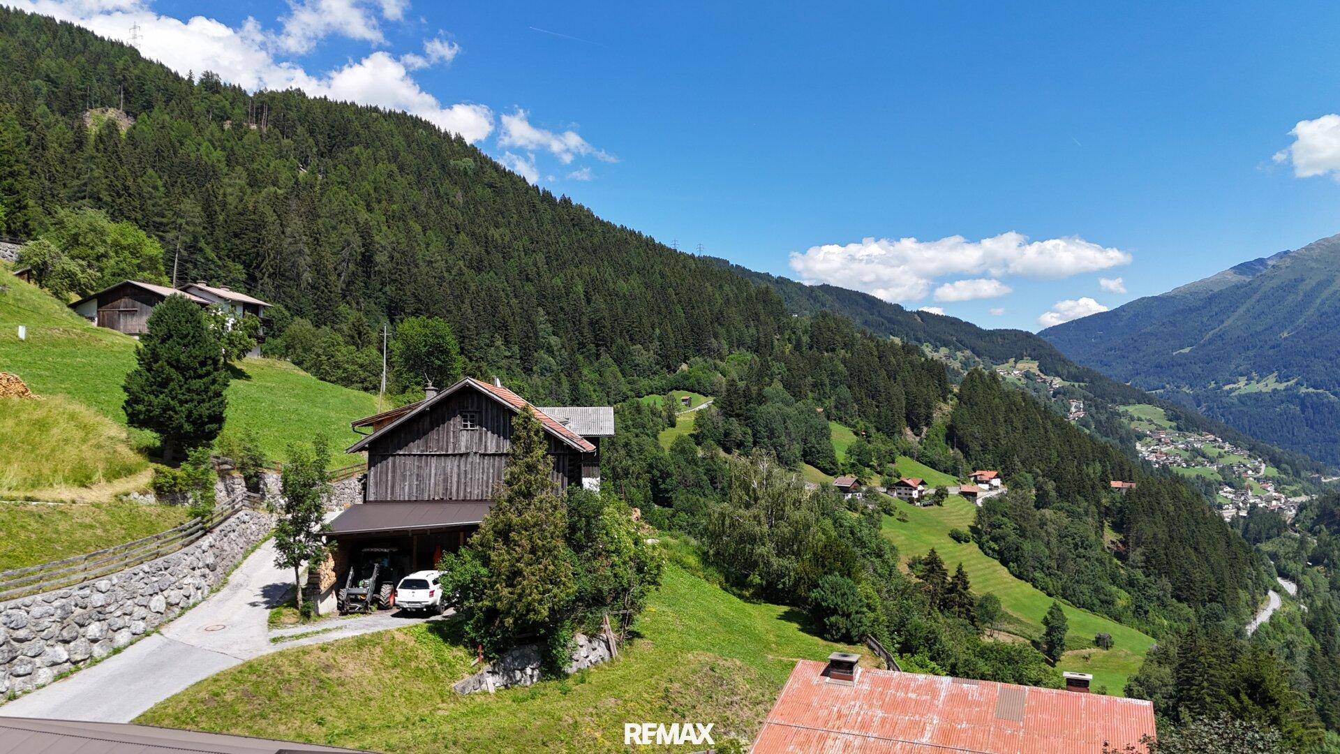 Traditionelles Holzhaus auf einem grünen Hang mit Blick auf die umliegenden Berge und das Tal.