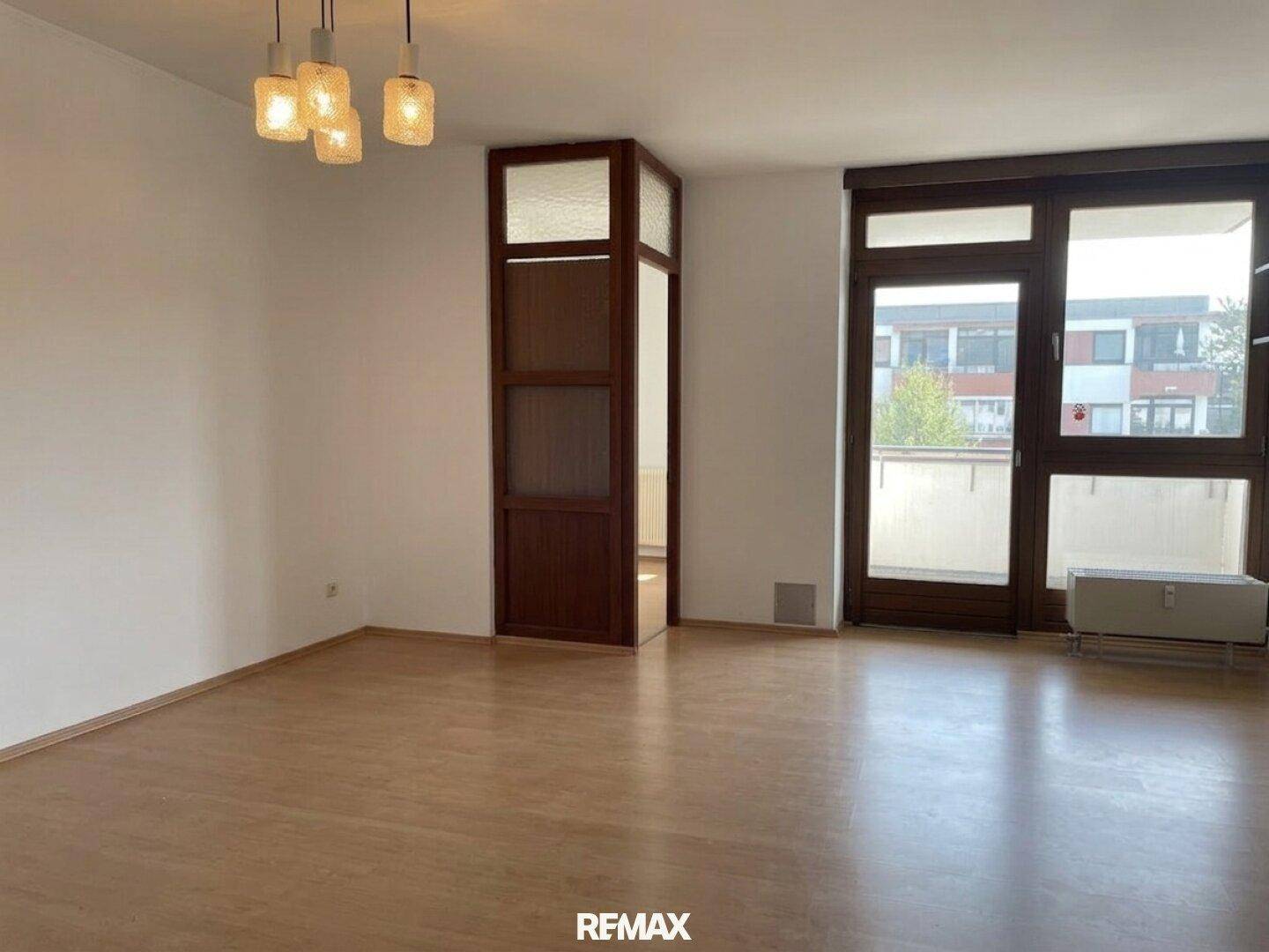 Helles, leeres Wohnzimmer mit Laminatboden und großen Fenstern, die viel Tageslicht hereinlassen.