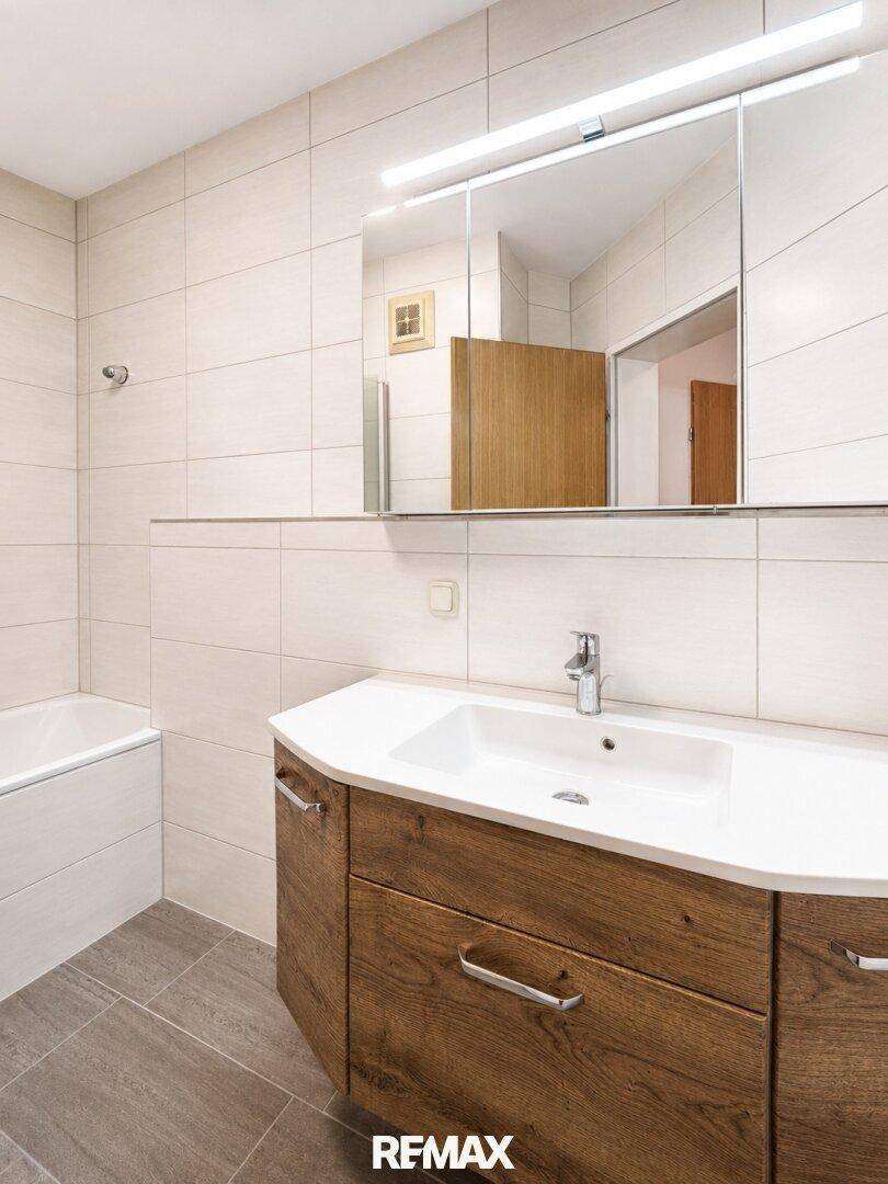Modernes Badezimmer mit Holzwaschtisch, Spiegelschrank und hellen Fliesen an Wänden und Boden.