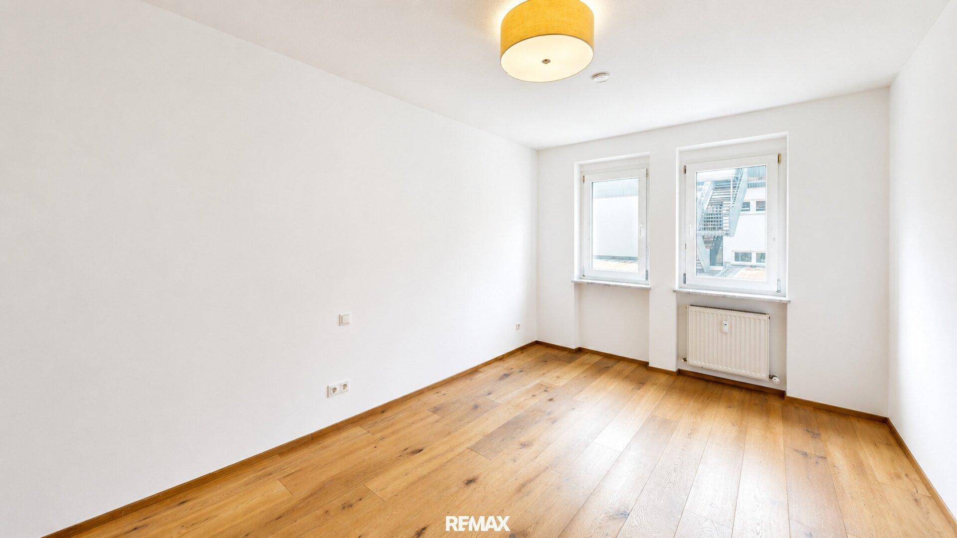 Leeres Schlafzimmer mit hellem Parkettboden, weißen Wänden und zwei Fenstern für Tageslicht.