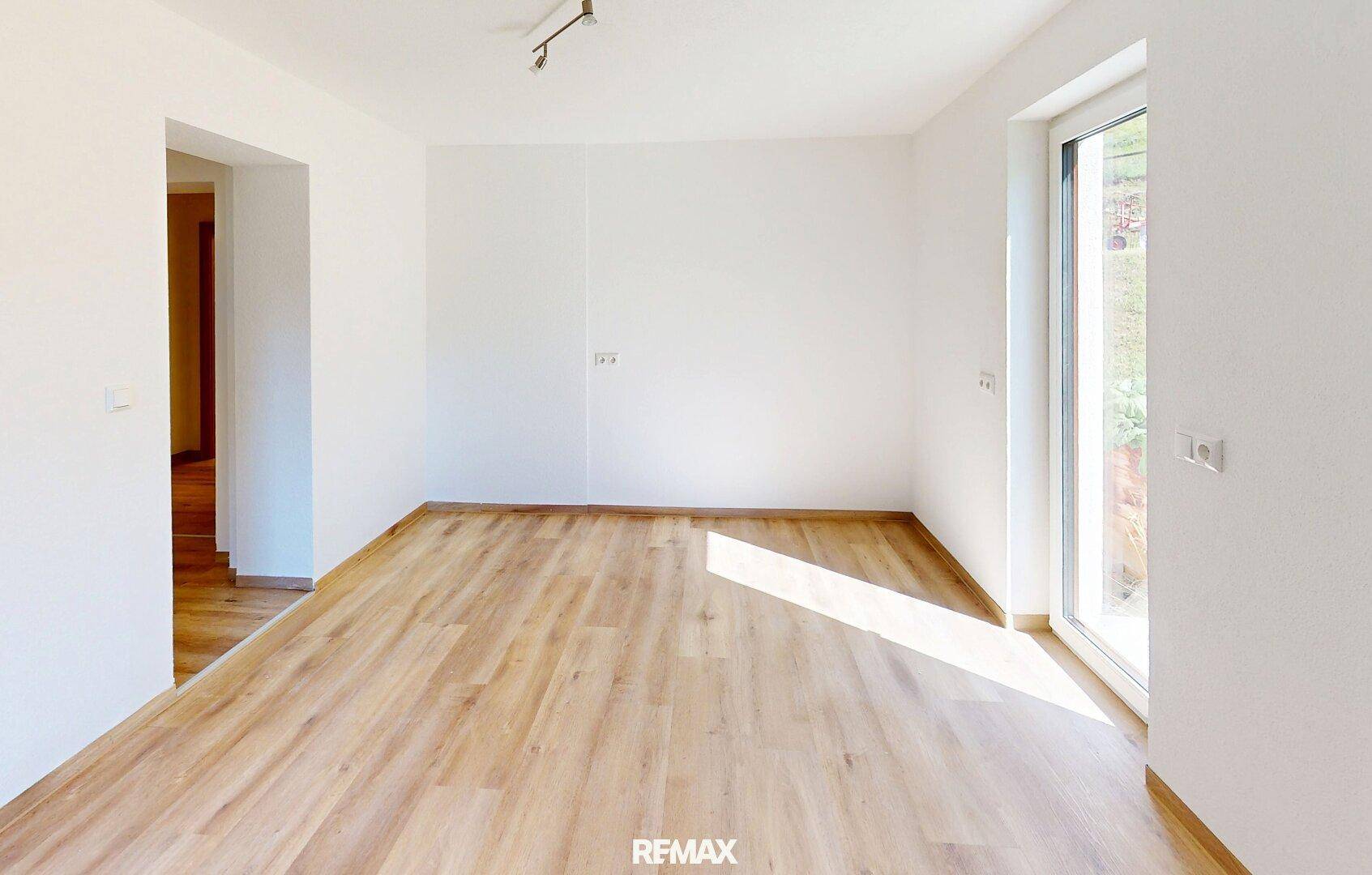 Leerer Raum mit hellem Laminatboden und großem Fenster, das viel Tageslicht hereinlässt.