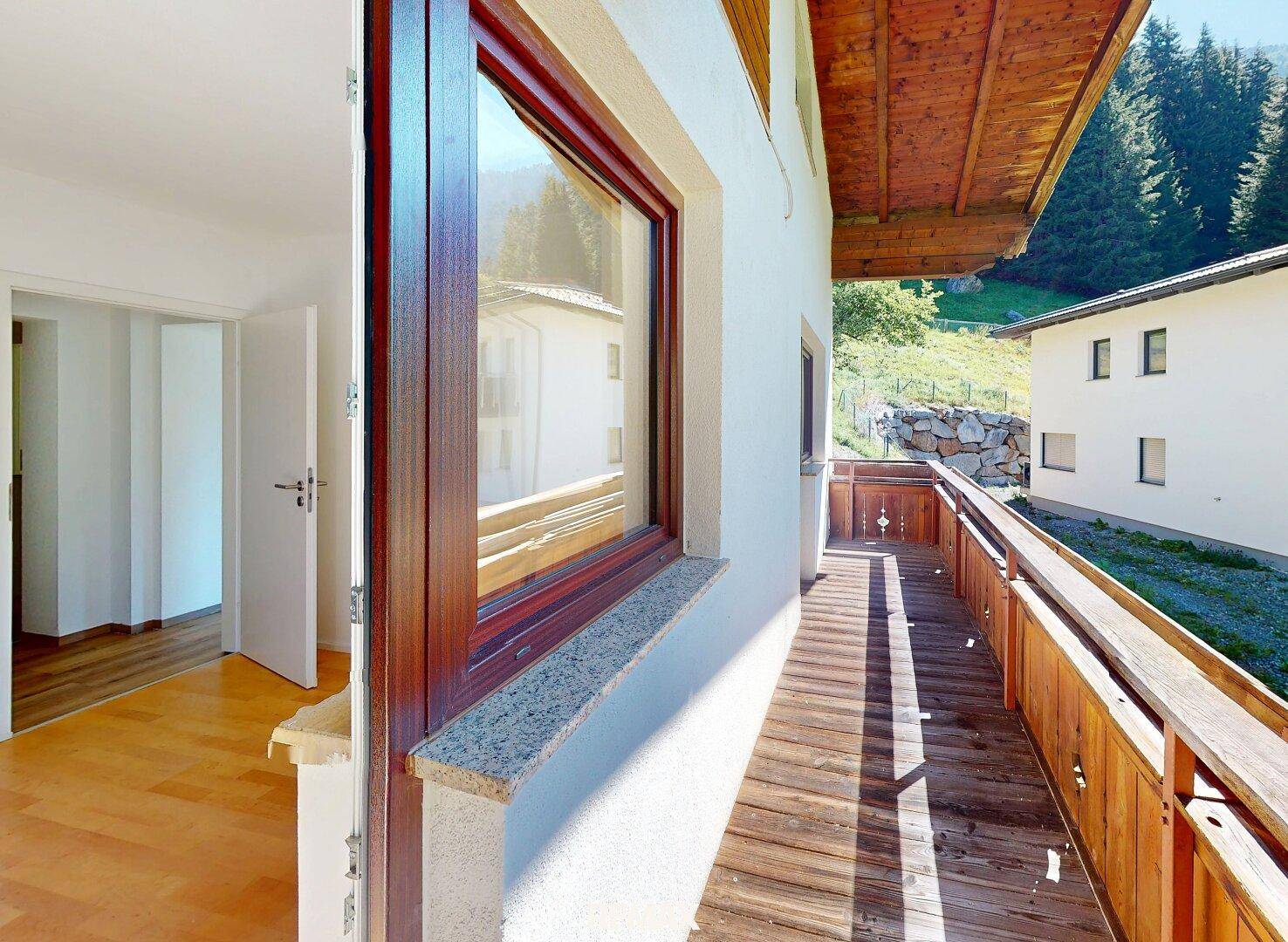Langer Holzbalkon mit Blick auf die grüne Hügellandschaft und angrenzenden Innenraum.