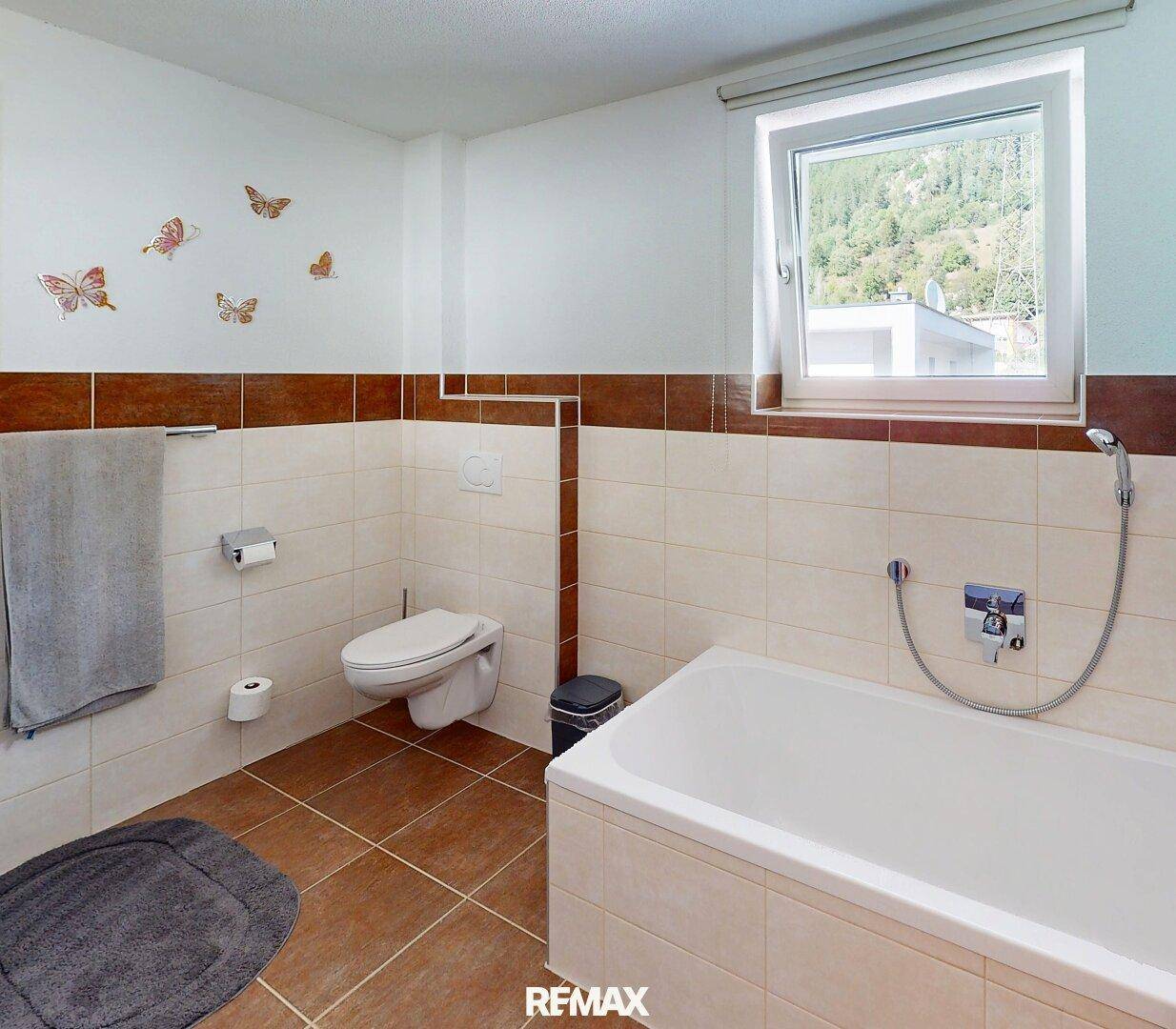 Badezimmer mit Badewanne, WC und braun-beigen Fliesen, sowie einem Fenster.