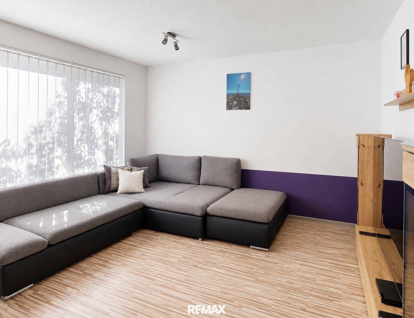 Helles Wohnzimmer mit großem Ecksofa, Laminatboden und vertikalen Jalousien am Fenster.