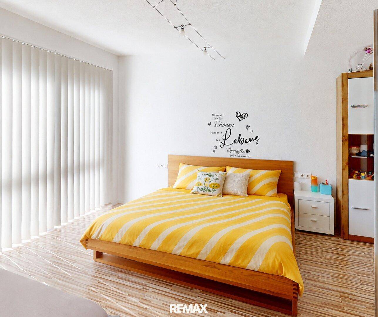 Schlafzimmer mit Holzbett, gelb-weiß gestreifter Bettwäsche und großem Fenster.