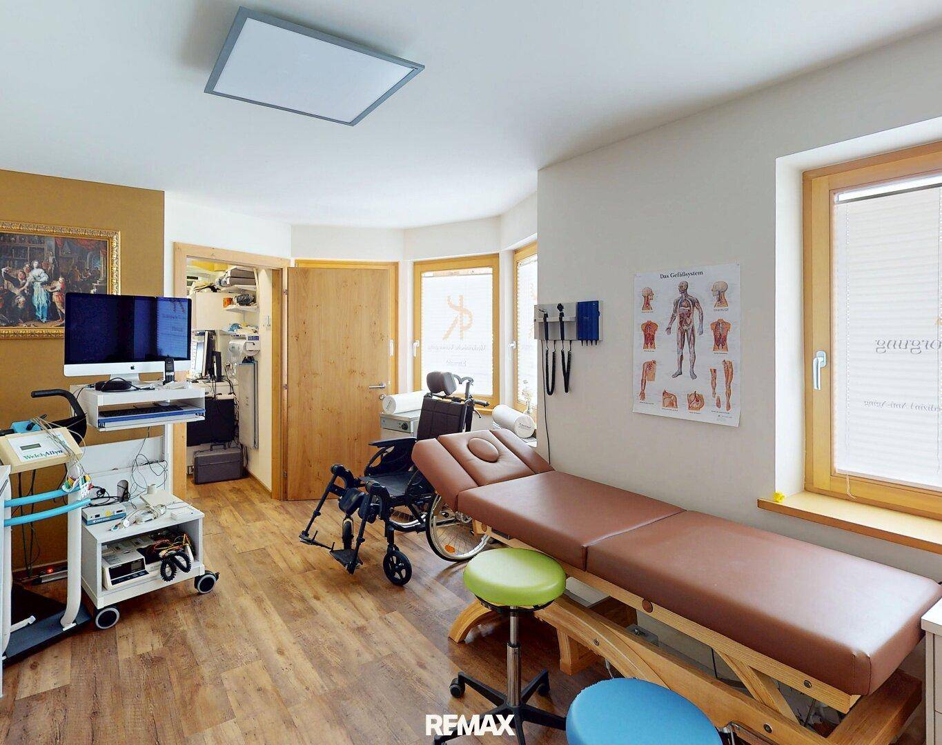Behandlungsraum mit Untersuchungsliege, Rollstuhl und medizinischen Diagrammen an der Wand.