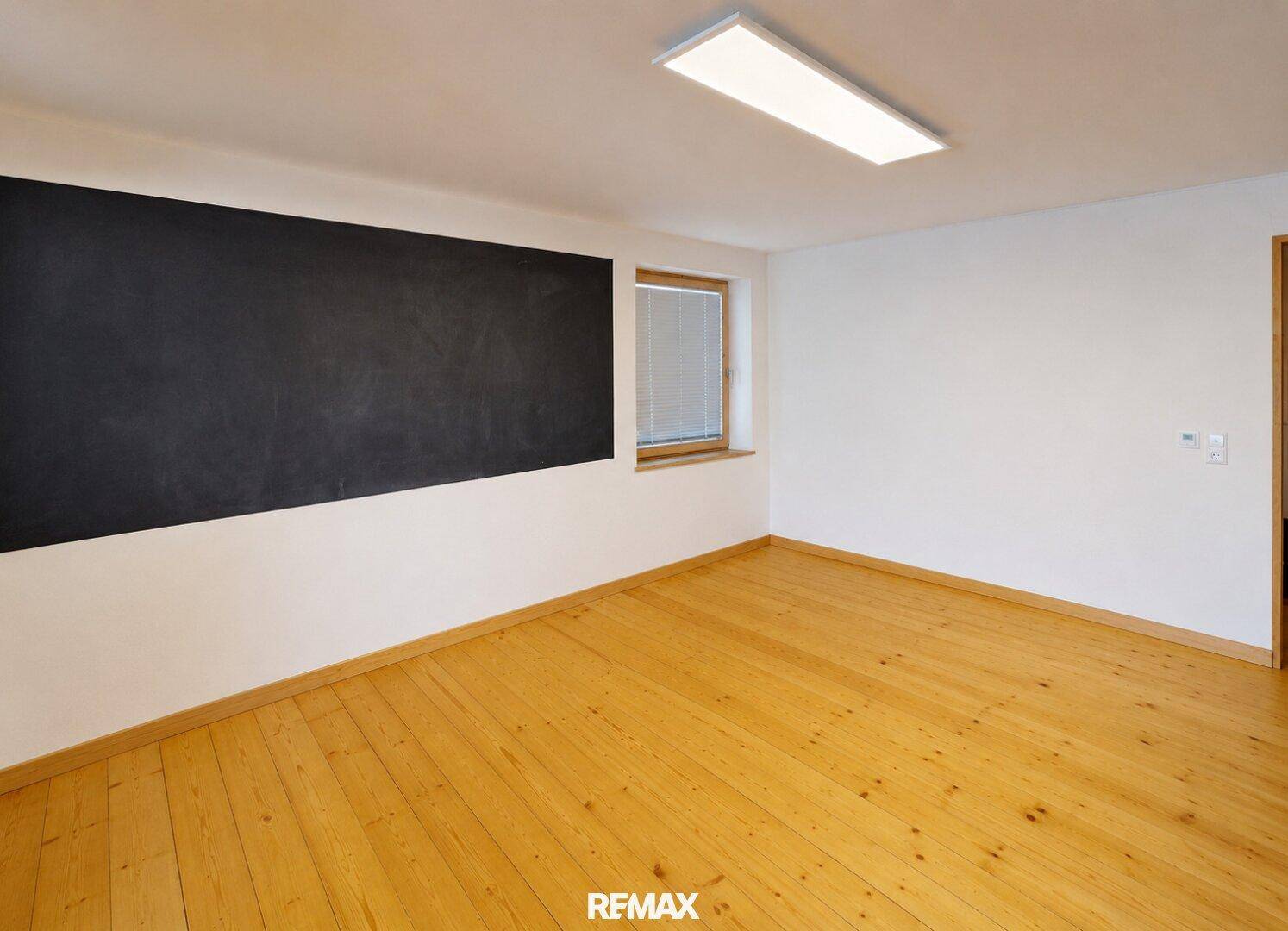 Heller Raum mit Holzboden, Fenster und einer schwarzen Wandtafel.