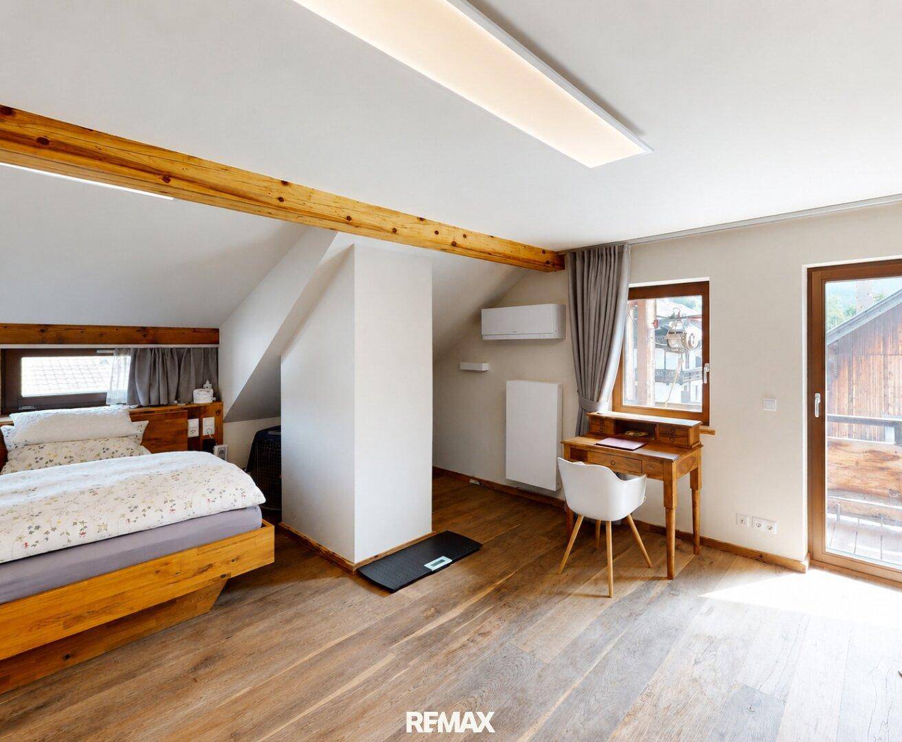 Helles Schlafzimmer mit Holzbalken, Schreibtisch und Zugang zum Balkon.