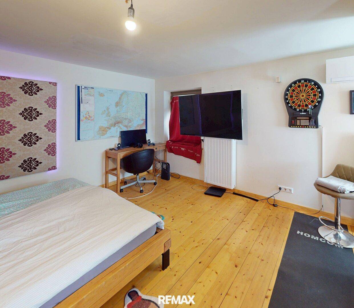 Schlafzimmer mit Holzboden, Schreibtisch, Fernseher und Dartscheibe.