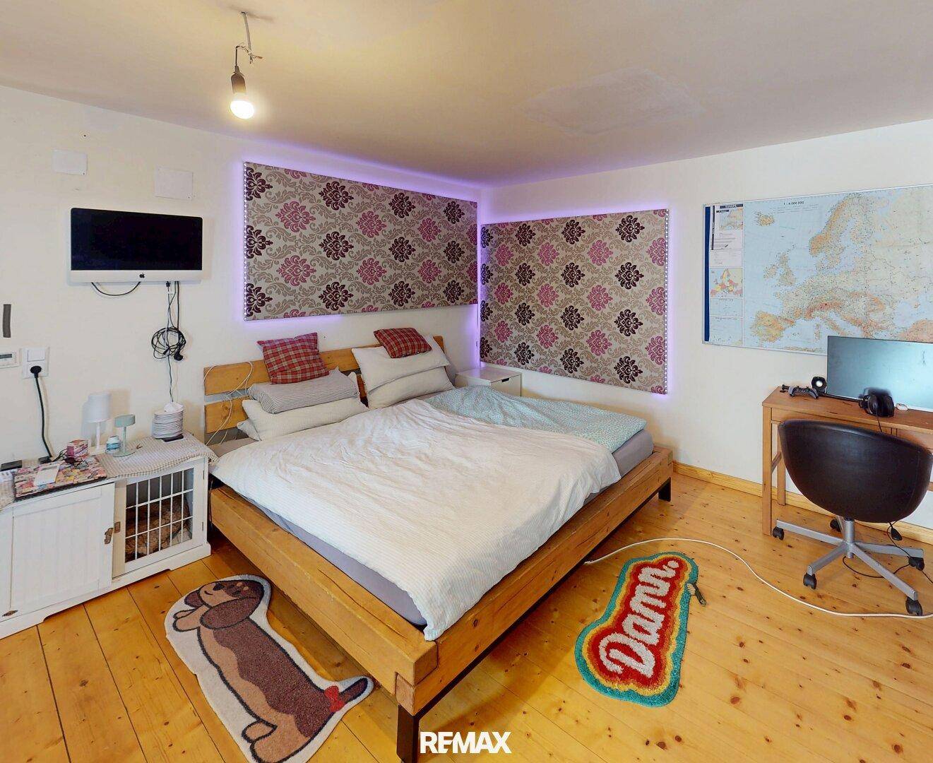 Schlafzimmer mit Holzboden, Doppelbett, Schreibtisch und farbiger Beleuchtung.