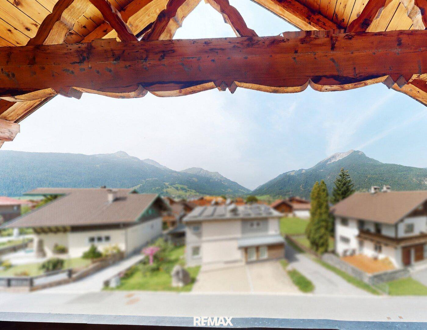 Panoramablick auf die umliegende Berglandschaft von einem überdachten Holzbalkon aus.