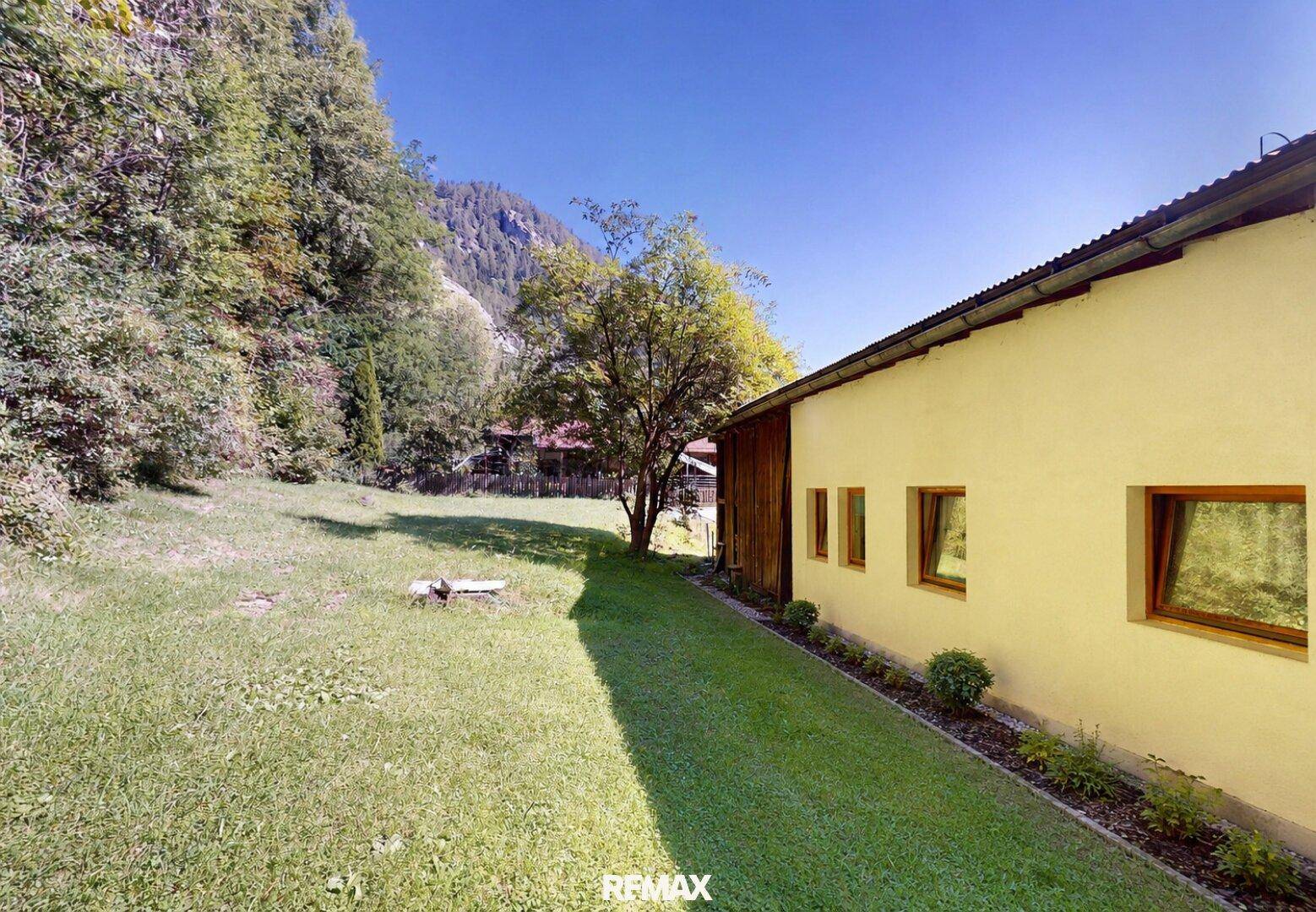 Seitenansicht des Hauses mit grünem Rasen, Bäumen und einer felsigen Berglandschaft im Hintergrund.