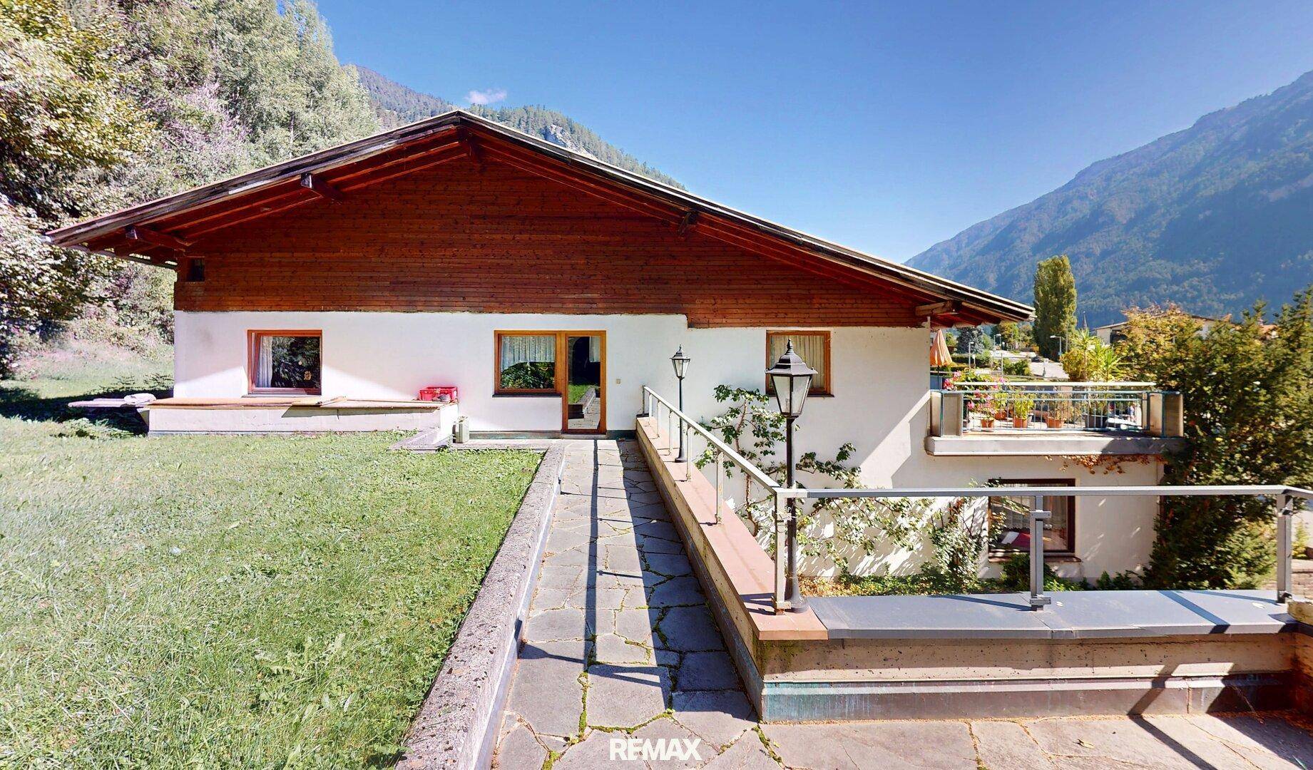 Traditionelles Haus mit Holzelementen und gepflegtem Rasen vor einer beeindruckenden Bergkulisse.
