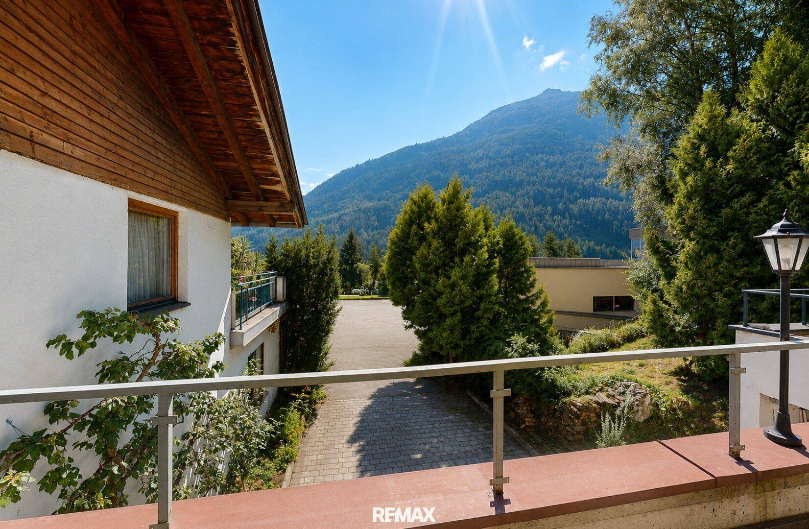 Blick von einem Balkon auf eine gepflasterte Fläche, Bäume und die umliegende Berglandschaft.