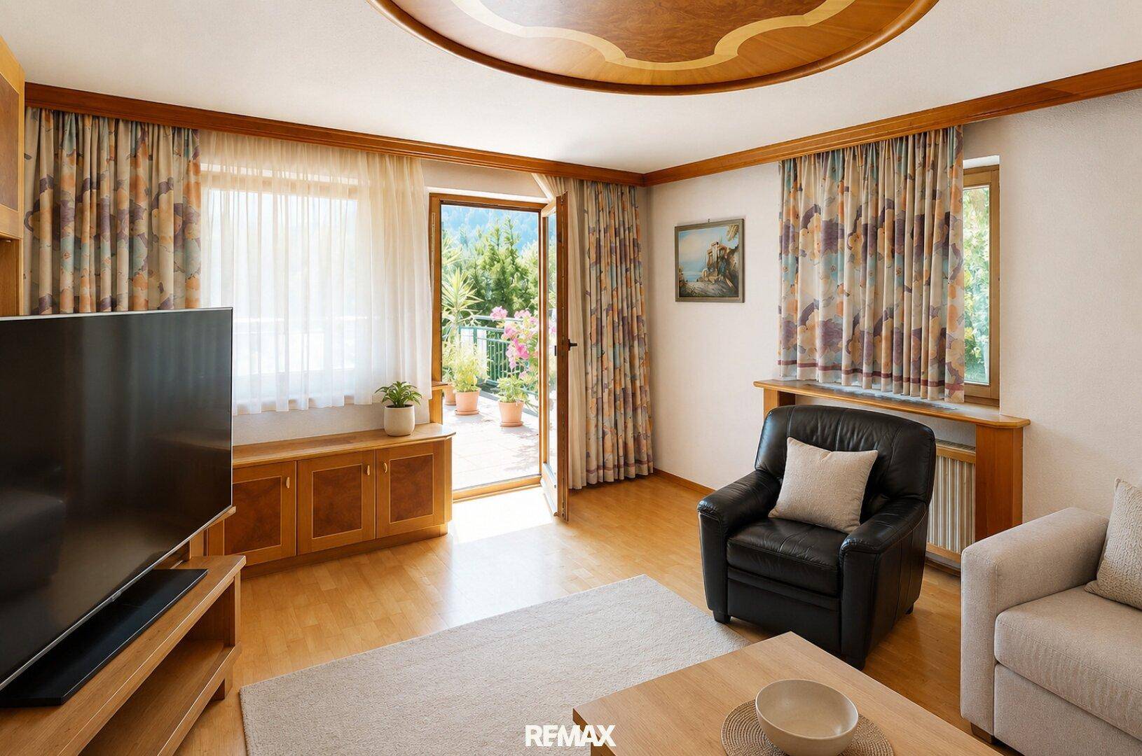 Gemütliches Wohnzimmer mit Sofa, Sessel, Fernseher und Zugang zu einem Balkon mit Bergblick.