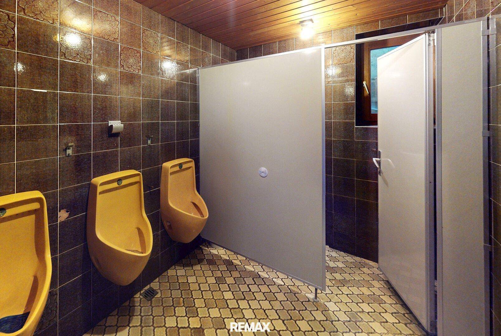 Öffentliche Toilette mit mehreren Urinalen und braunen Fliesen an Wänden und Boden.