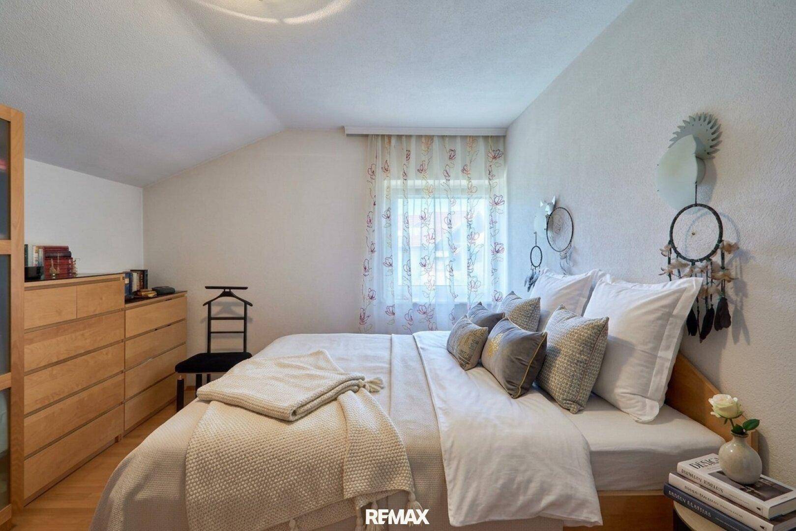 Helles Schlafzimmer mit Holzboden, Dachschräge und Fenster mit Vorhängen.