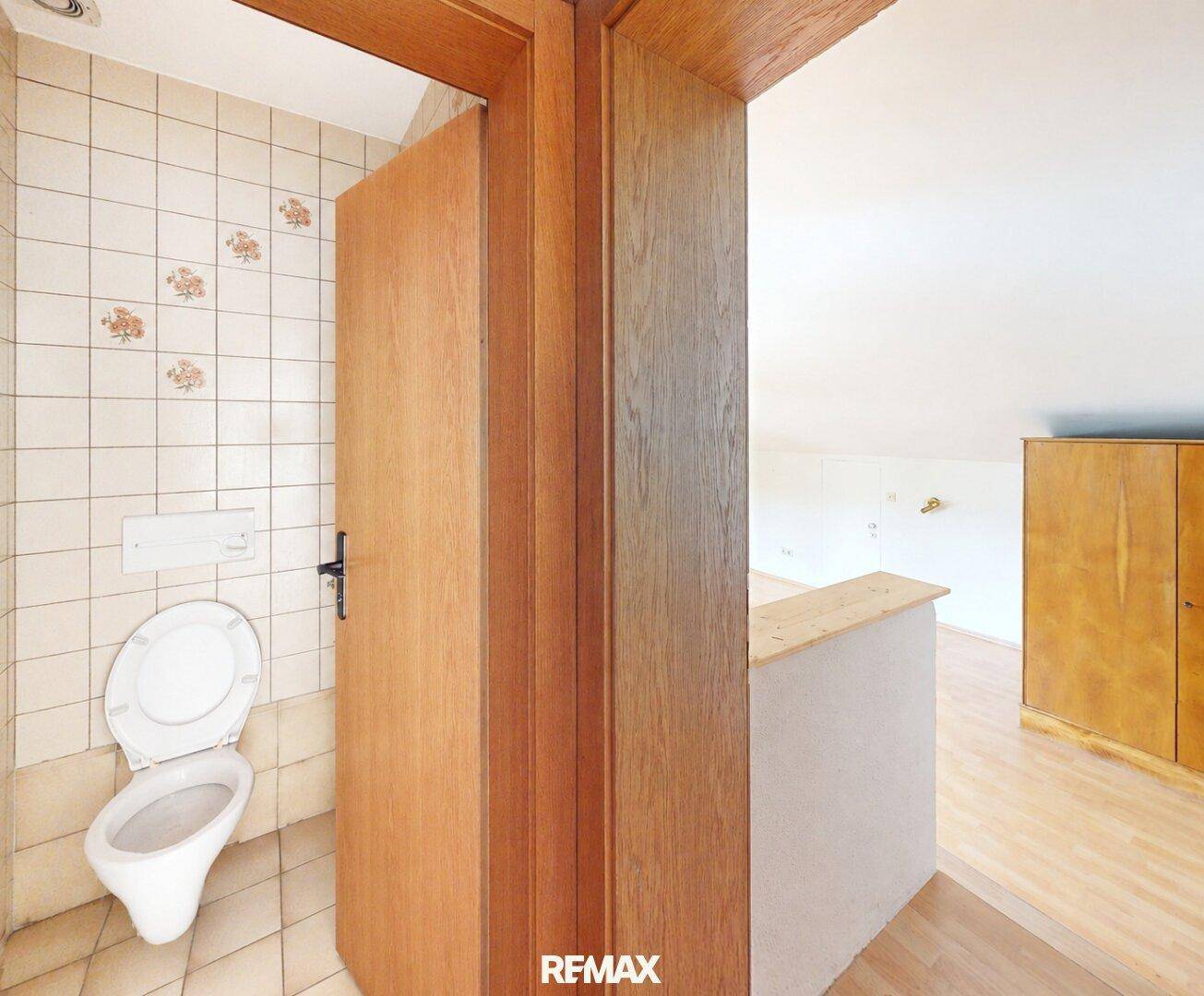 Separate Toilette mit beigefarbenen Fliesen und Holztür, angrenzend an einen weiteren Wohnbereich.