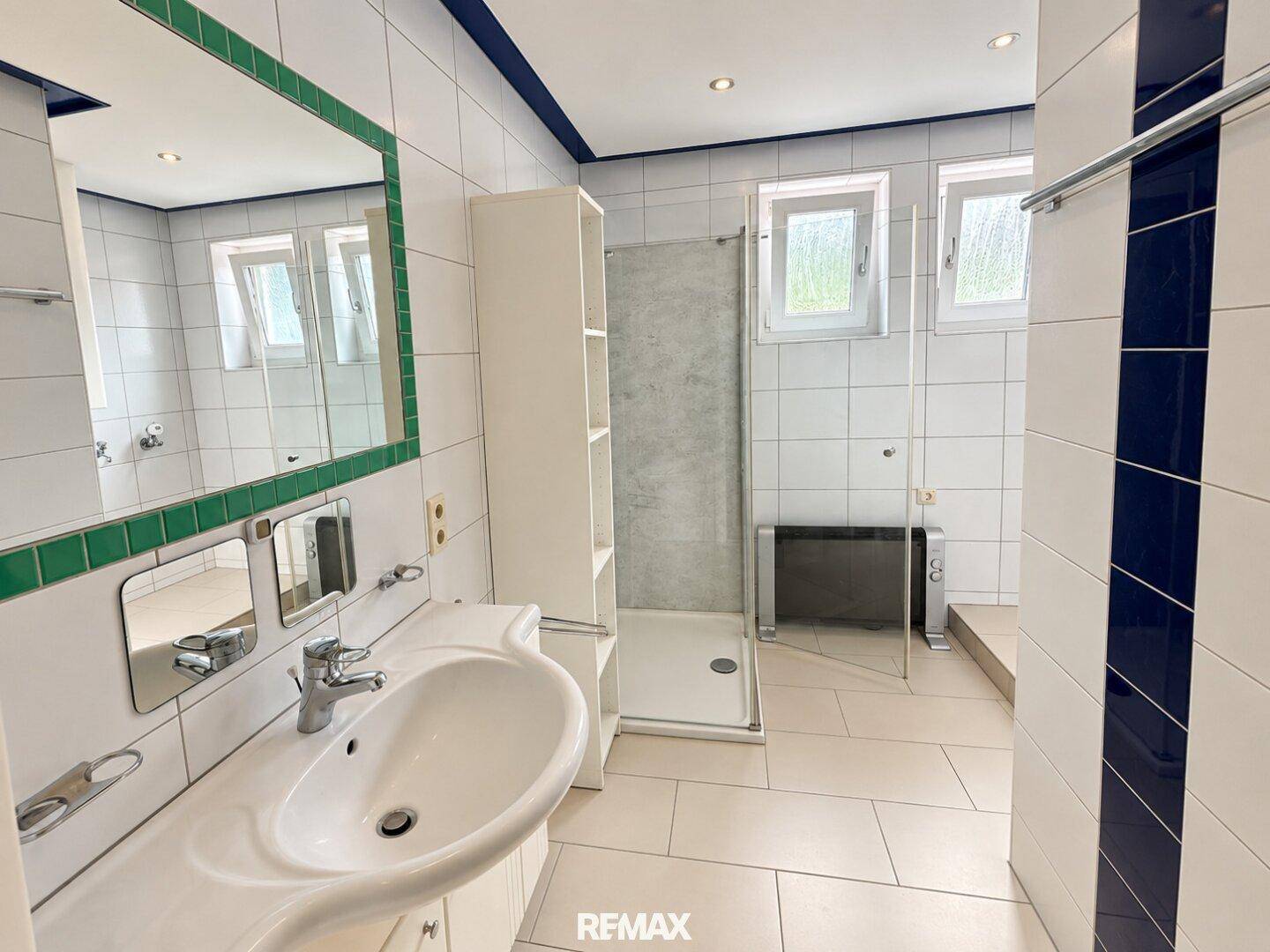 Badezimmer mit Dusche, Waschbecken und Fenster, ausgestattet mit weißen Fliesen und farbigen Akzenten.