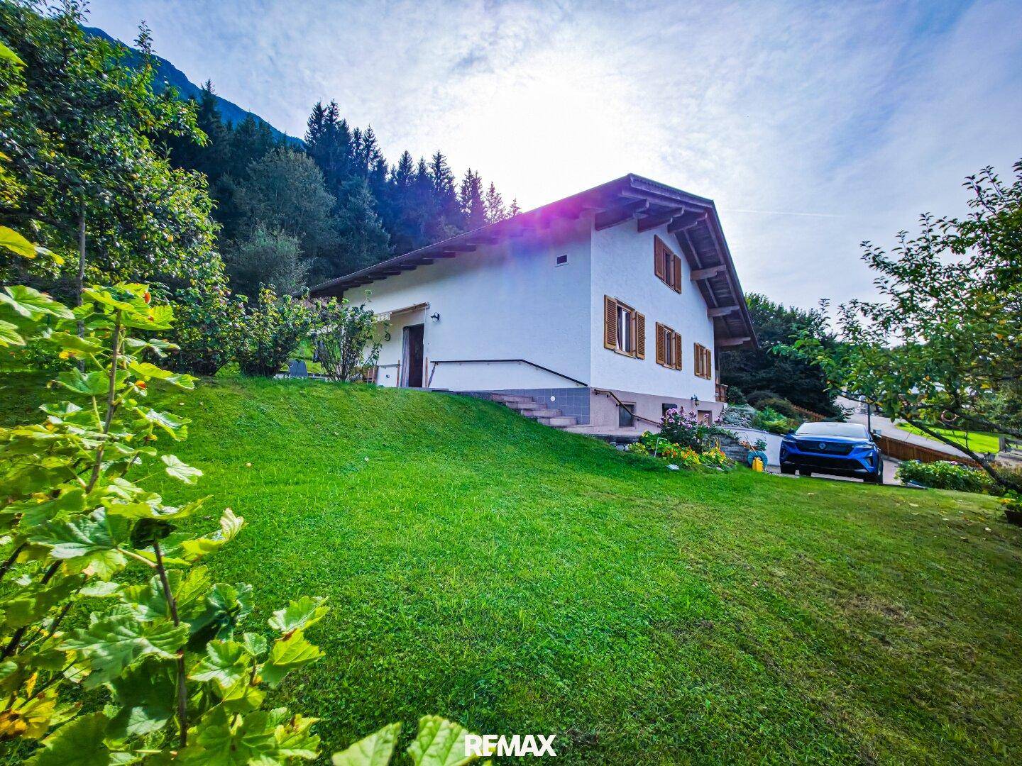 Weißes Einfamilienhaus mit Satteldach und großem, grünem Garten in Hanglage mit Bergblick.