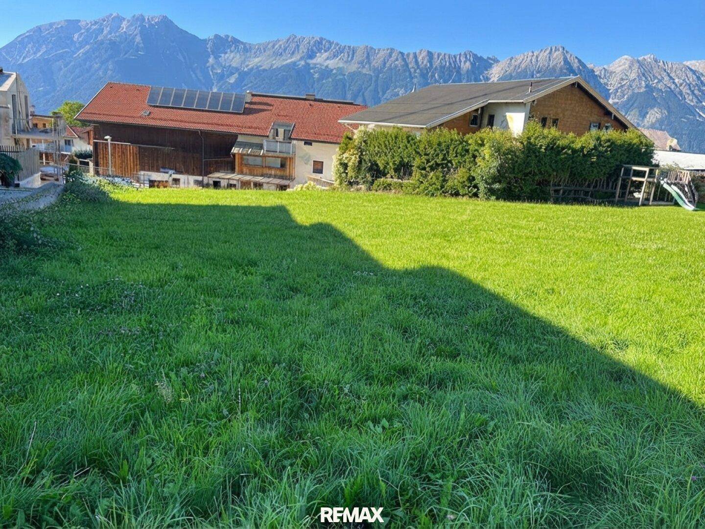 Sonnige Grünfläche mit Häusern, einem Spielgerät und beeindruckendem Alpenpanorama.