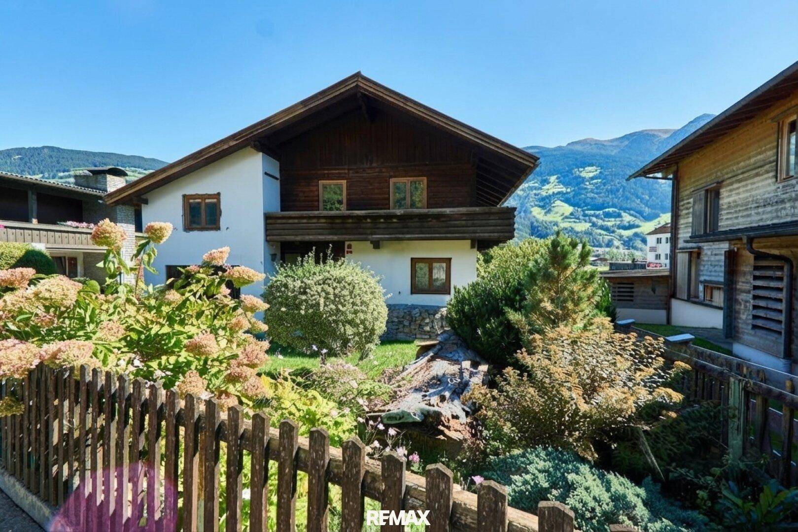 Vorderansicht eines charmanten Hauses im alpinen Stil mit Holzbalkon, Garten und Bergpanorama.