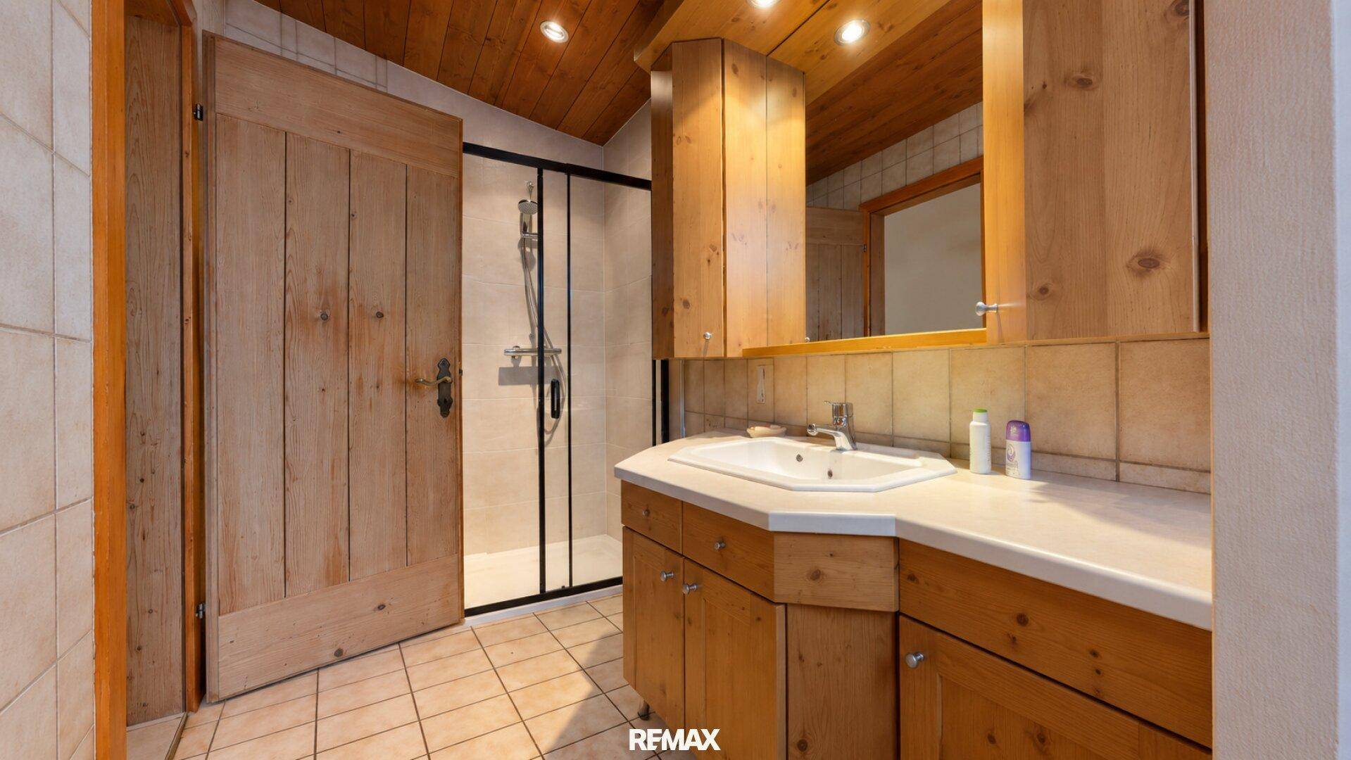 Badezimmer mit Dusche, Holzwaschtisch und Dachschräge, gefliest in hellen Tönen.