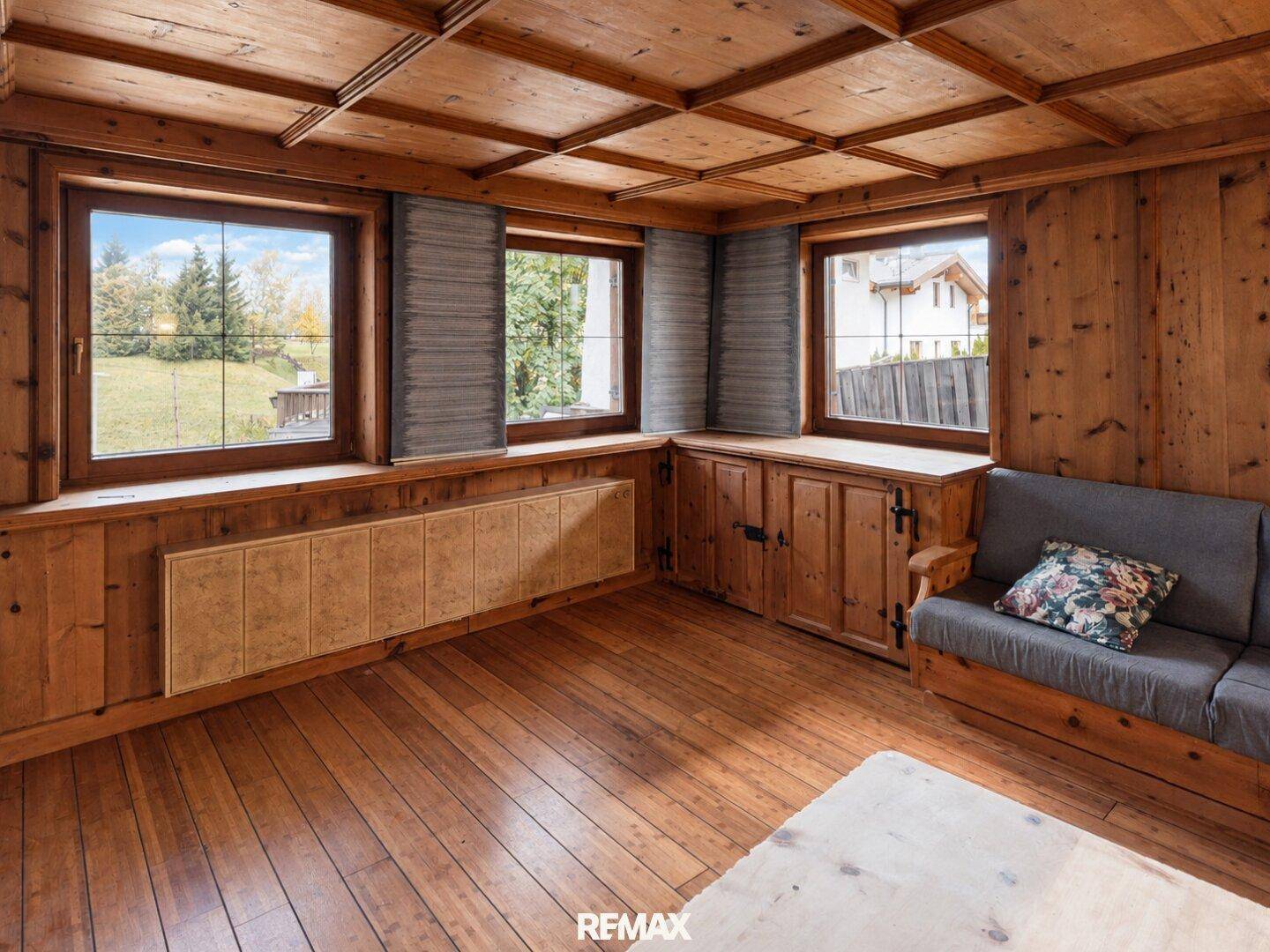 Rustikales Wohnzimmer mit Holztäfelung, Holzboden und Blick ins Grüne durch mehrere Fenster.