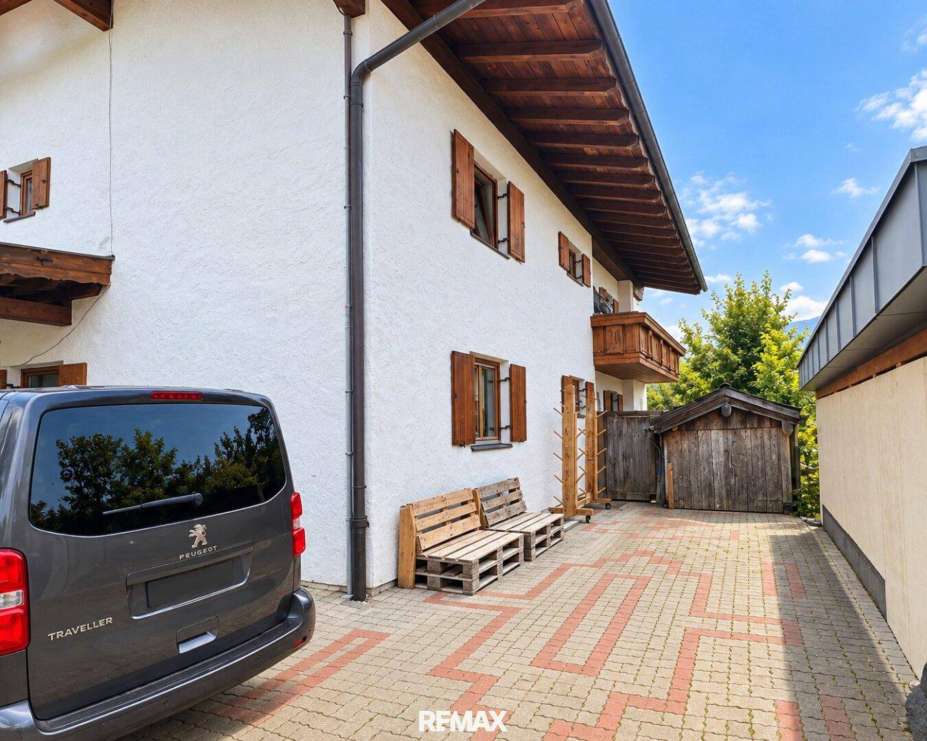 Seitenansicht des Hauses mit gepflasterter Fläche, geparktem Auto und einem kleinen Holzschuppen.