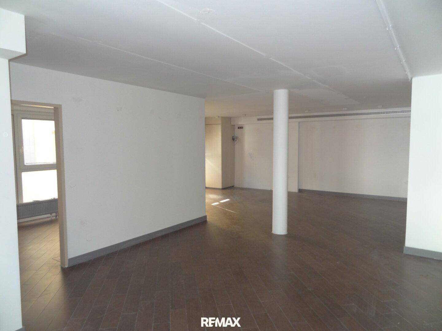 Leerer Gewerberaum mit dunklem Laminatboden, weißen Wänden und einem Fensterbereich, bereit zur Gestaltung.