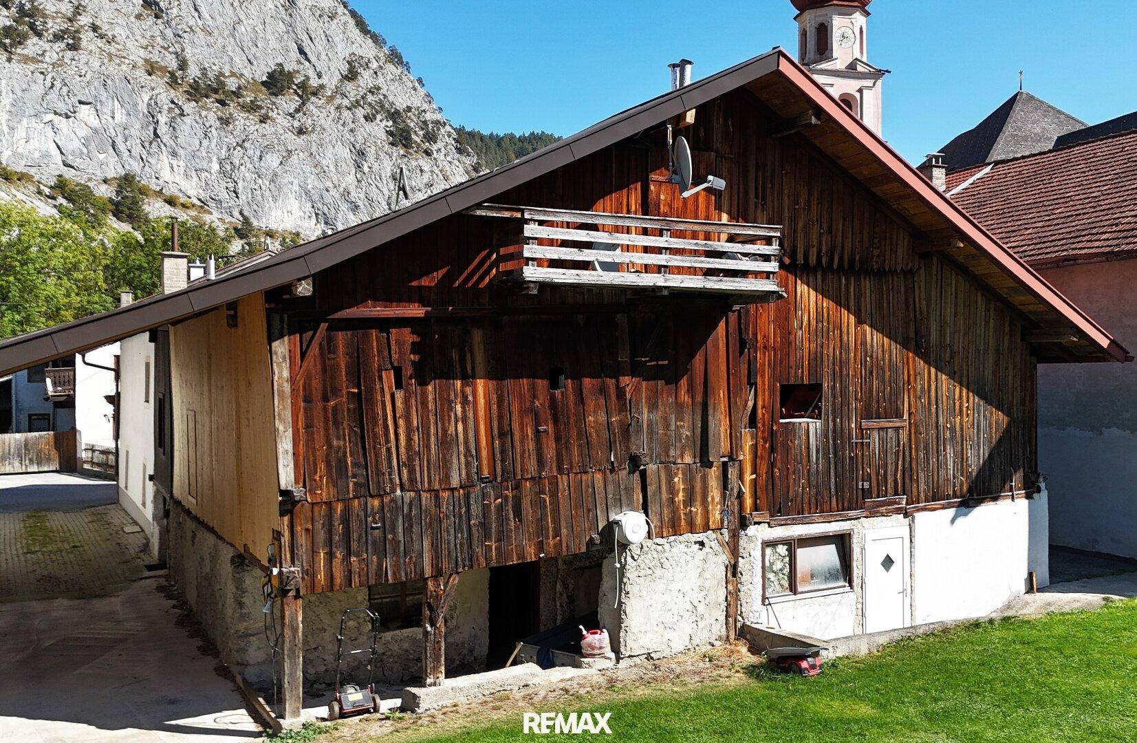 Rustikales Gebäude mit Holzwänden und einem kleinen Balkon, umgeben von grüner Landschaft und Bergen.
