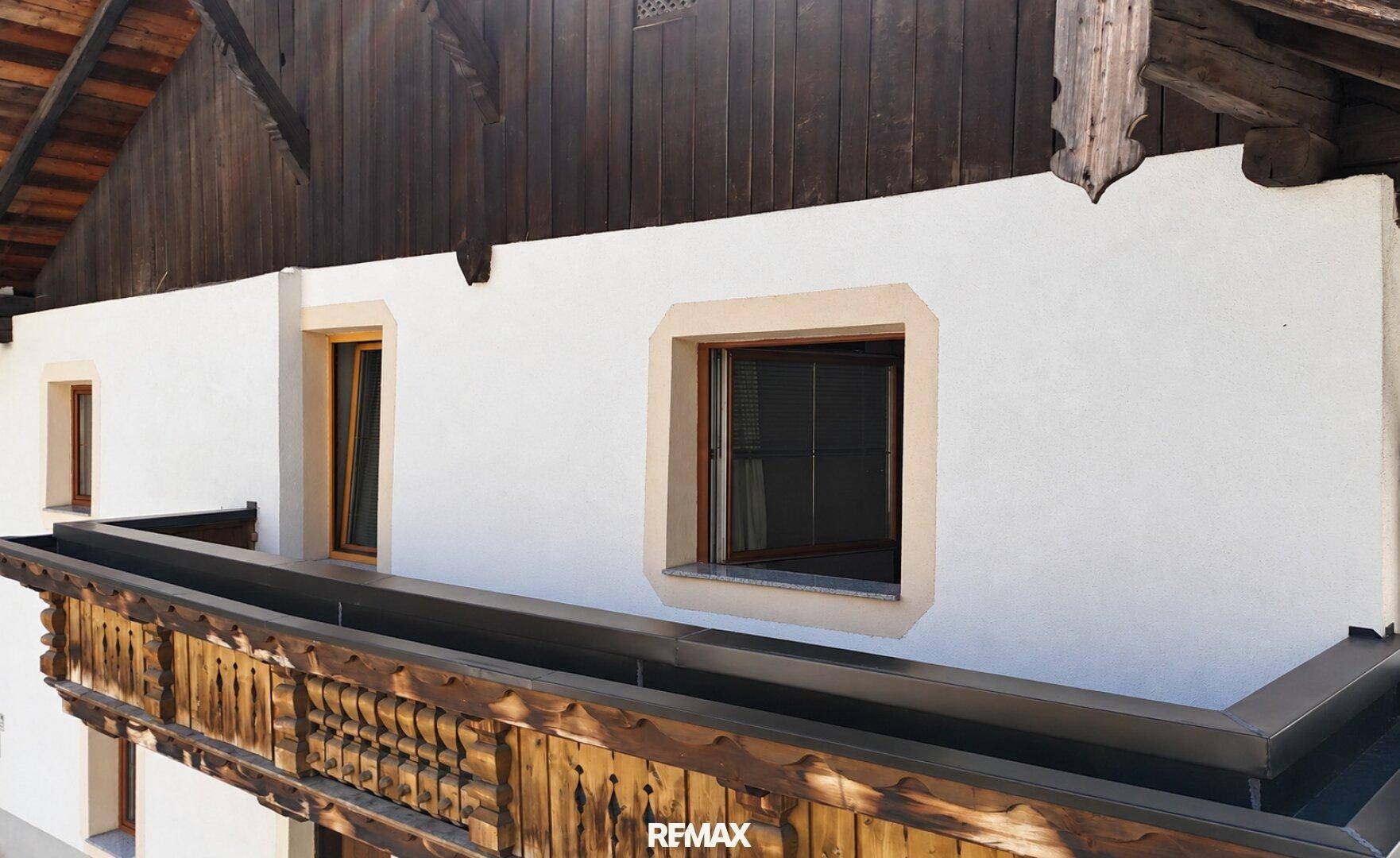 Traditioneller Holzbalkon mit kunstvollen Schnitzereien an einer weiß verputzten Fassade eines alten Gebäudes.