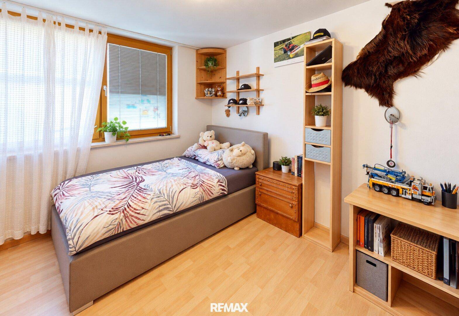 Kinderzimmer mit Einzelbett, Holzmöbeln und Fenster mit Jalousien.