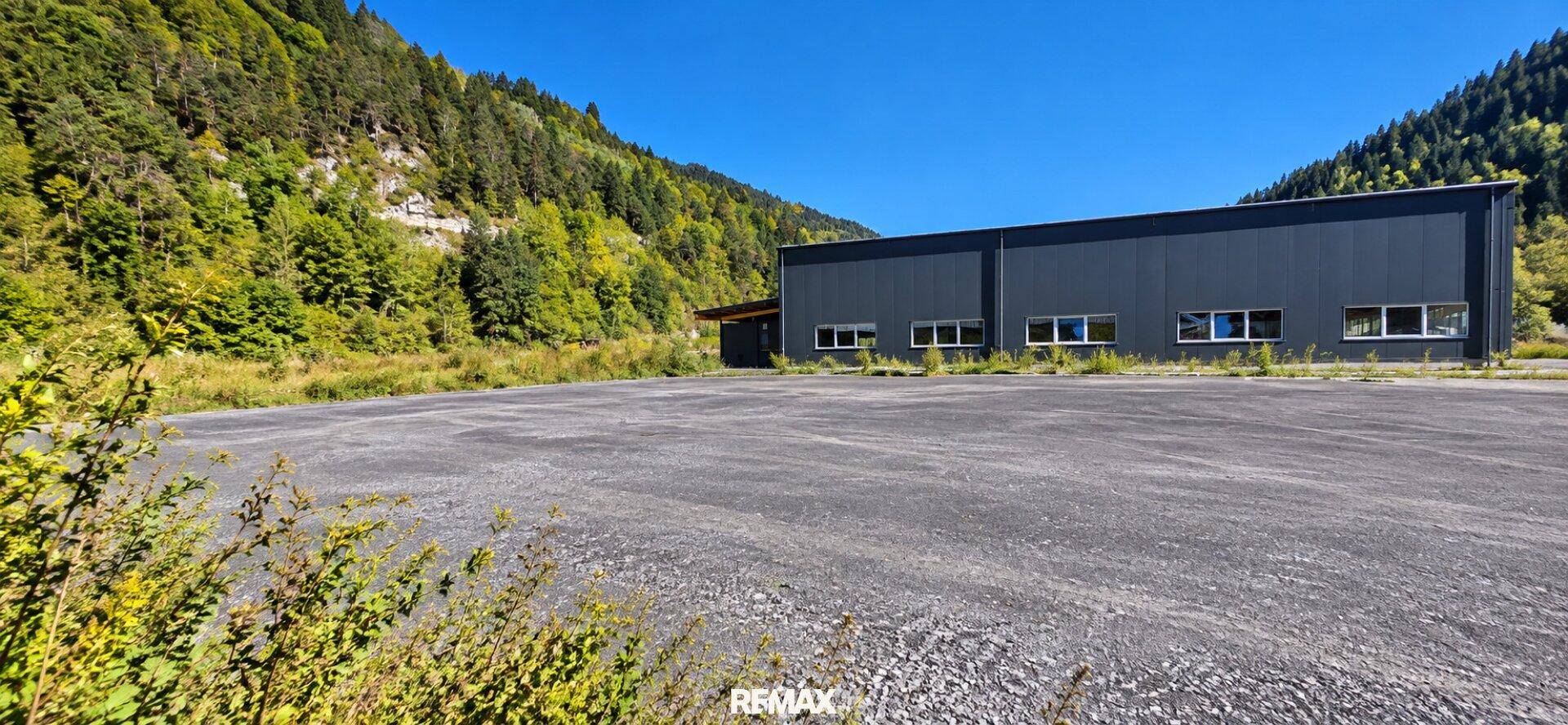 Weitläufige asphaltierte Fläche vor dem Industriegebäude mit bewaldeten Bergen im Hintergrund unter blauem Himmel.