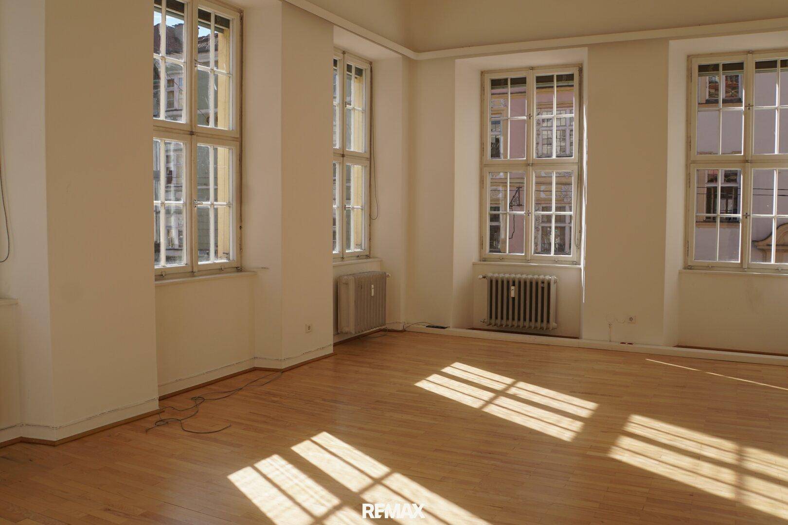 Leerer Raum mit hellem Holzboden, hohen Fenstern und Heizkörpern, durchflutet von natürlichem Sonnenlicht.