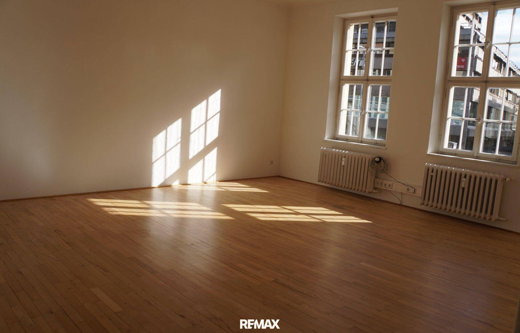 Heller, leerer Raum mit Holzboden und großen Fenstern, die viel Tageslicht hereinlassen und Schatten werfen.