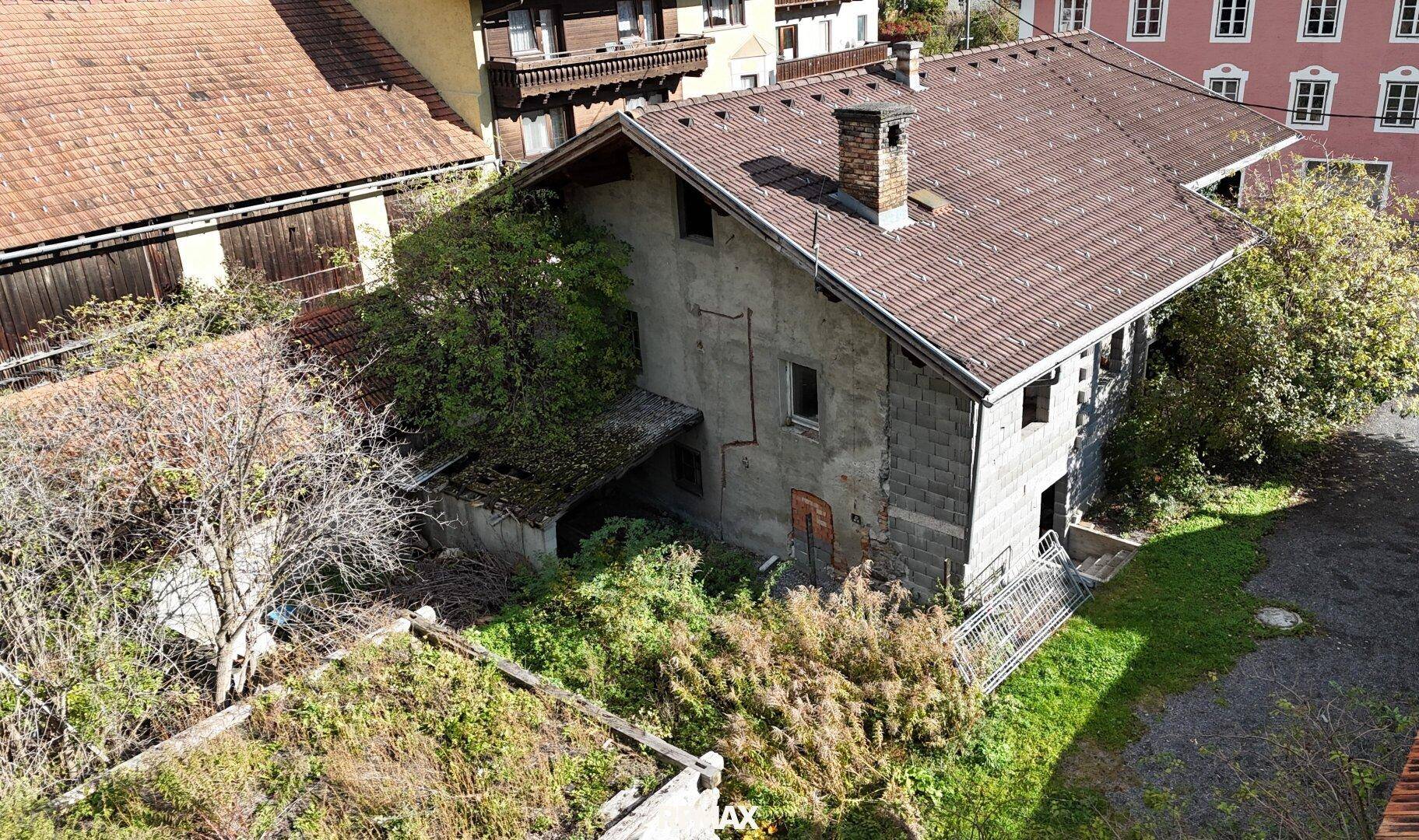 Seitenansicht eines älteren Hauses mit verwildertem Garten und Blick auf benachbarte Gebäude.