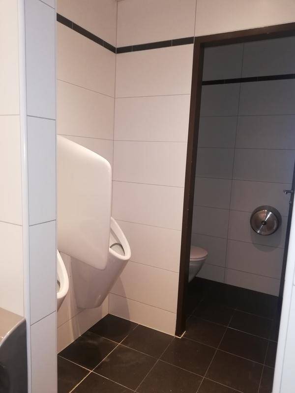 Gefliestes Badezimmer mit Urinalen und dunklem Fliesenboden, ein Durchgang führt zu einer weiteren Toilette.