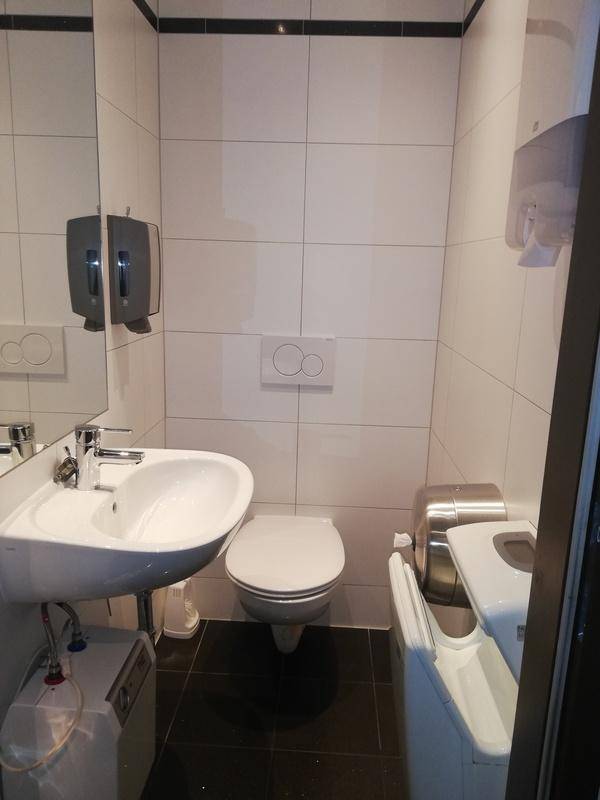 Kleines Badezimmer mit weißen Fliesen, einem Waschbecken, einer Toilette und einem Spiegel.