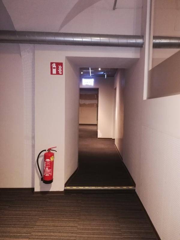 Langer Flur mit dunklem Teppichboden, sichtbaren Rohren an der Decke und einem Feuerlöscher an der Wand.