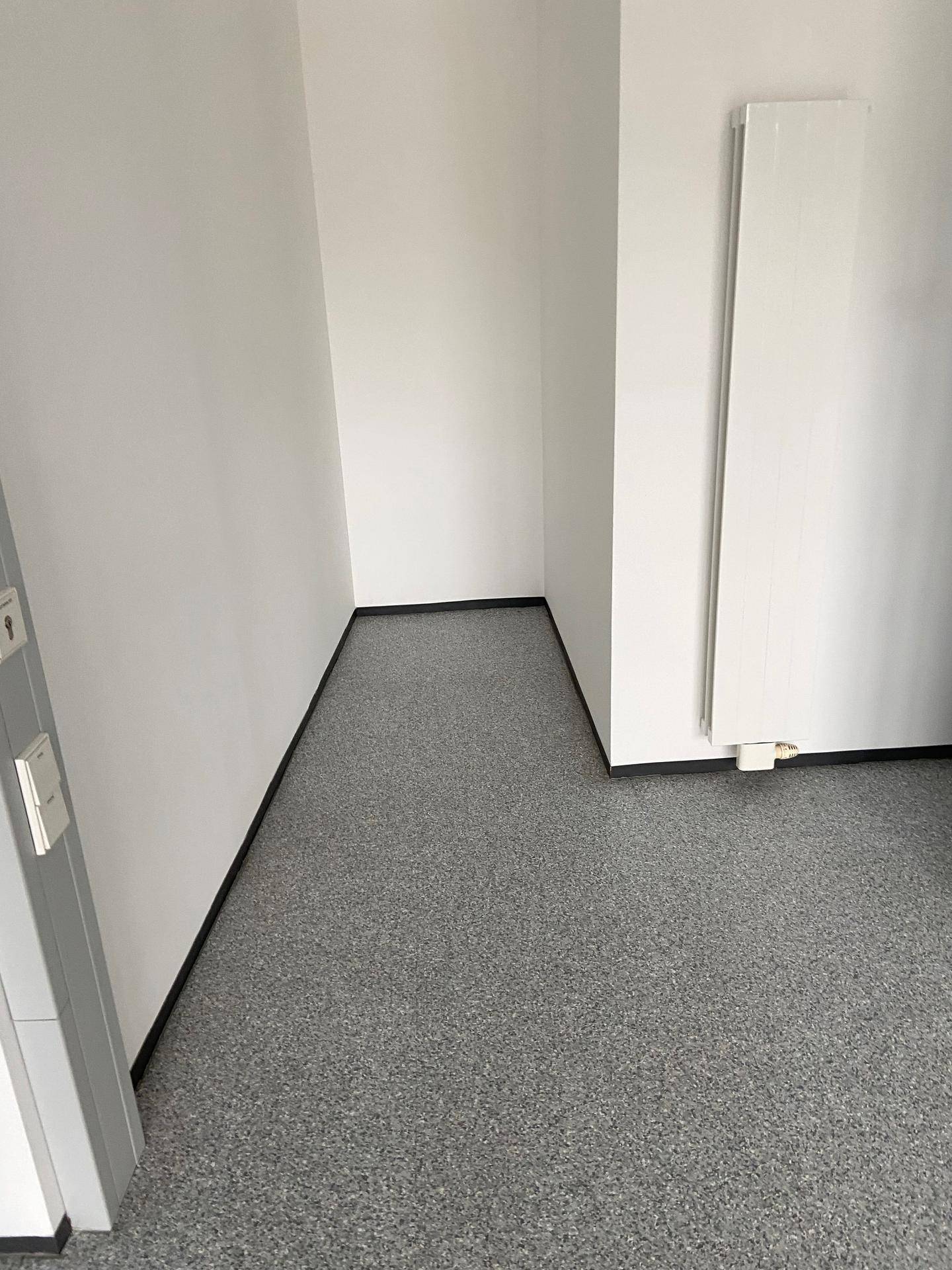 Ecke eines Raumes mit grauem Bodenbelag, weißen Wänden und einem Heizkörper.