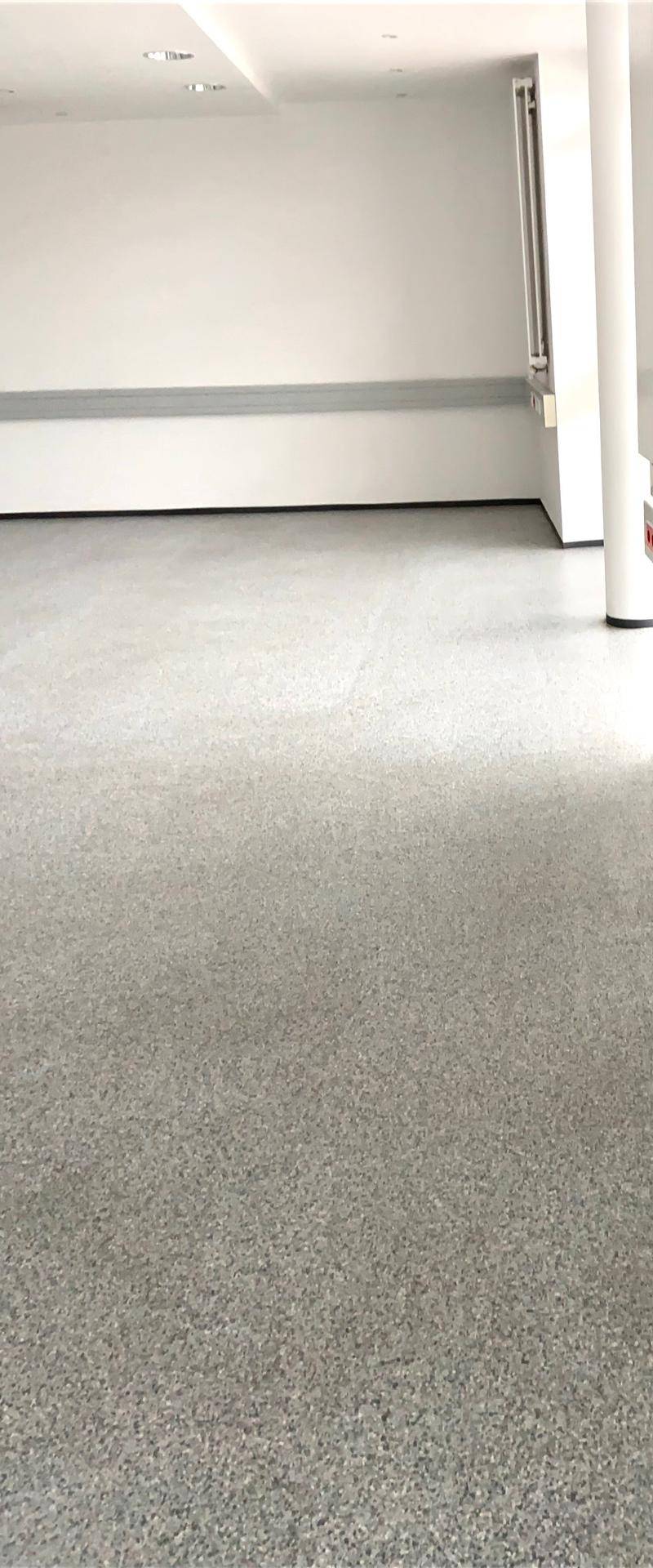 Detailansicht des grauen, gesprenkelten Bodenbelags und der weißen Wand in einem hellen Raum.