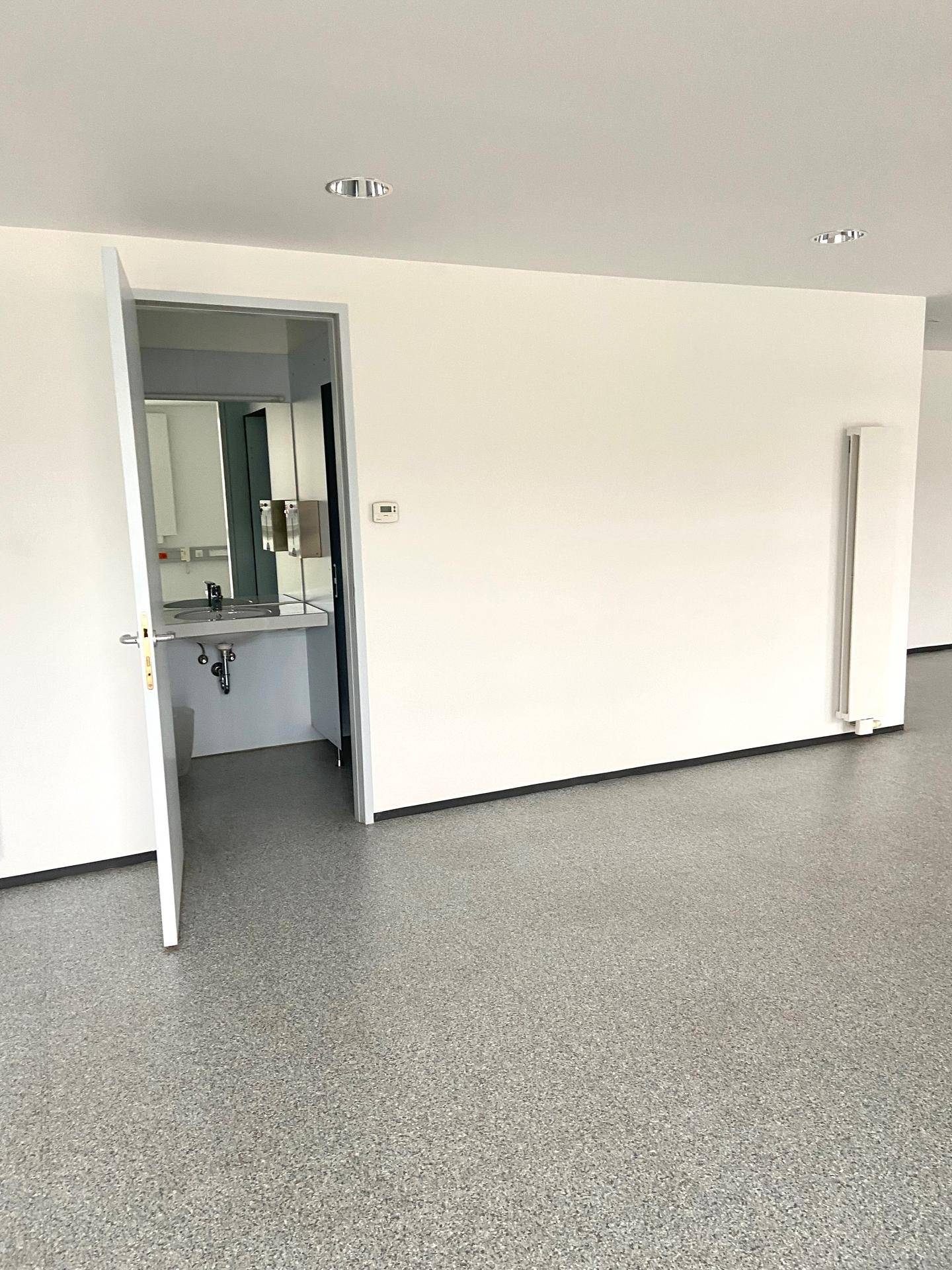 Heller, leerer Raum mit grauem Bodenbelag und offener Tür zu einem Waschbereich.