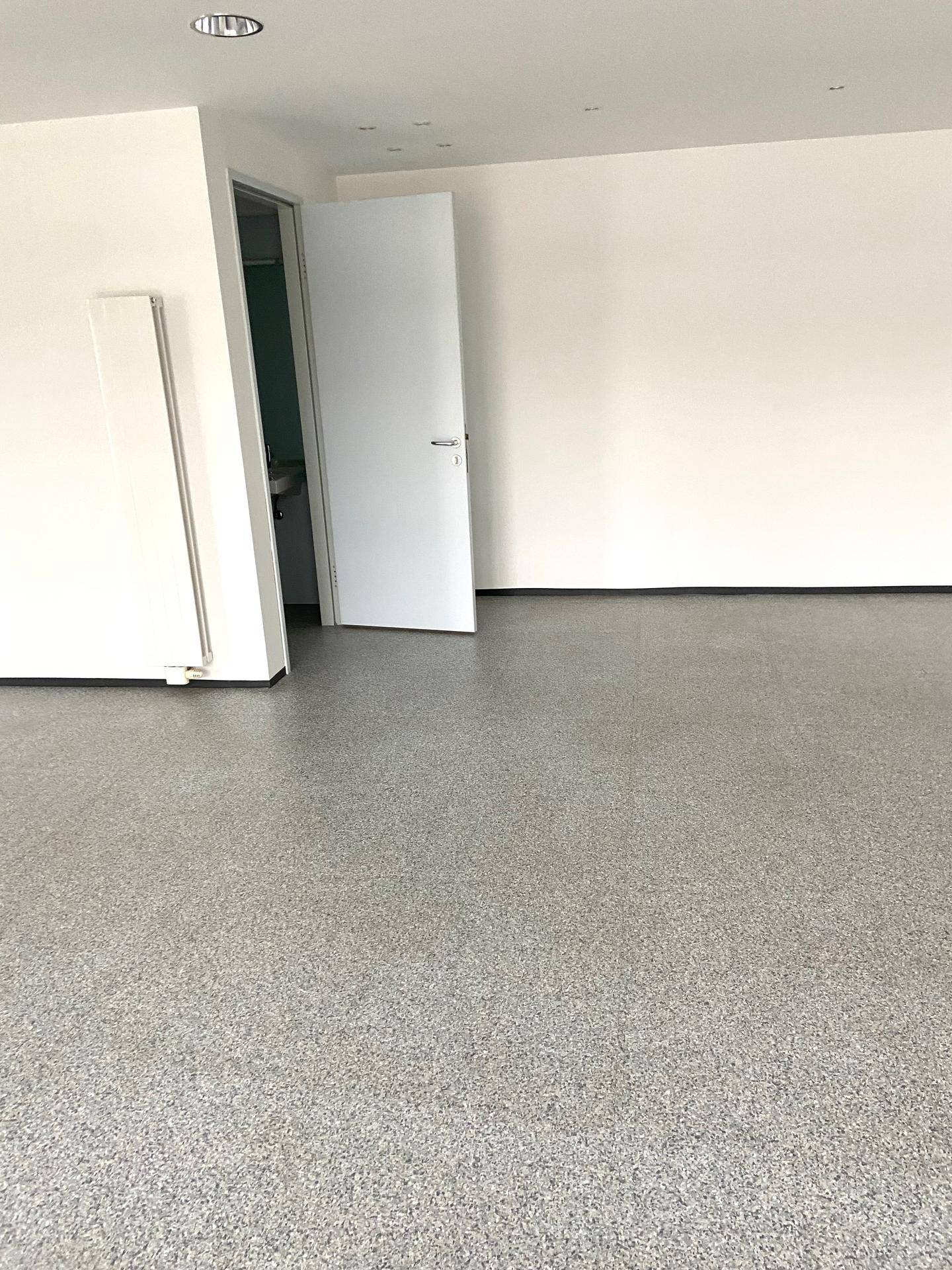 Leerer Raum mit grauem Bodenbelag und offener Tür zu einem Sanitärbereich, inklusive Heizkörper.