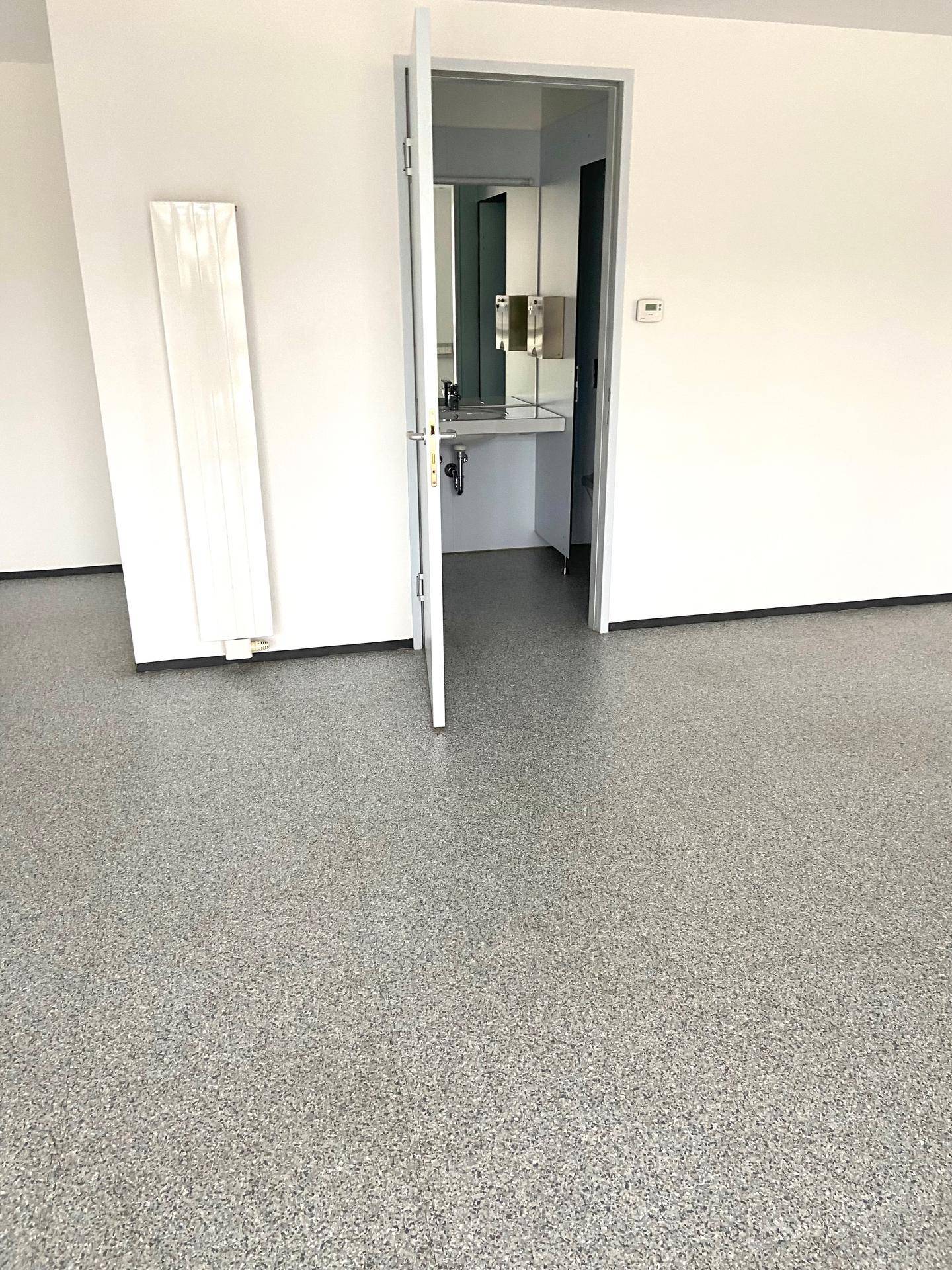 Geräumiger, leerer Raum mit grauem Bodenbelag und offener Tür zu einem Waschbereich.