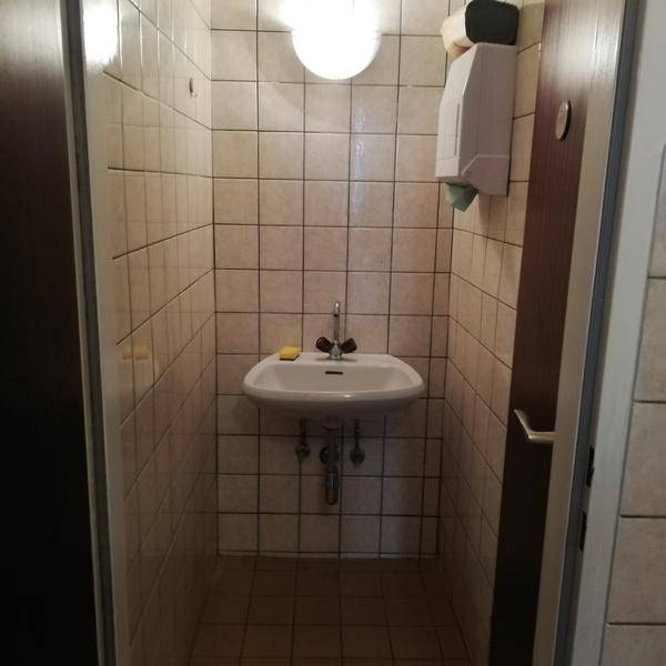 Kleiner gefliester Raum mit Waschbecken und Seifenspender, beleuchtet von einer Deckenlampe.
